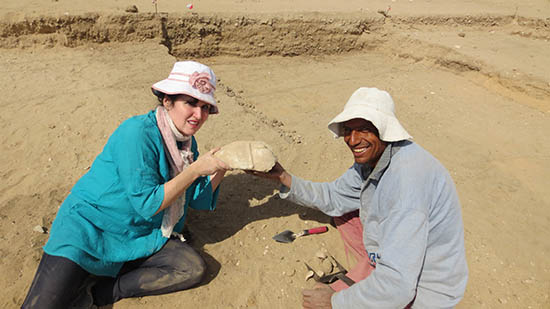 Laura and Mustafa crouch in the sand holding an artifact they have discovered.