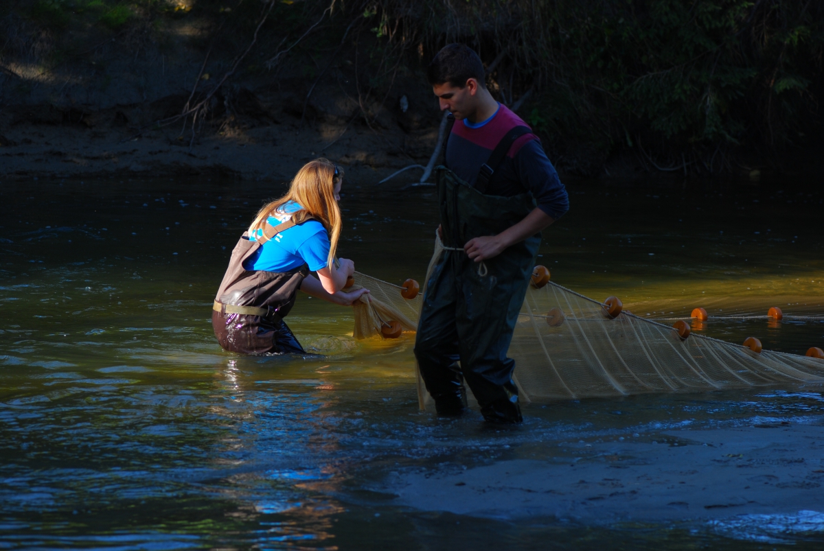 A woman and a man wearing hip waders with a net stand in a river