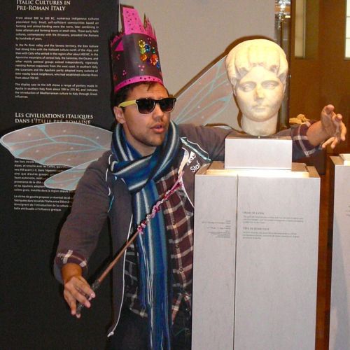 Kiron Mukherjee dressed as a fairy, posing with a roman bust on display in the Eaton Gallery of Rome.