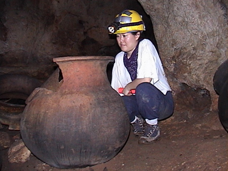 Kay crouches beside a large pot inside a dark cave.