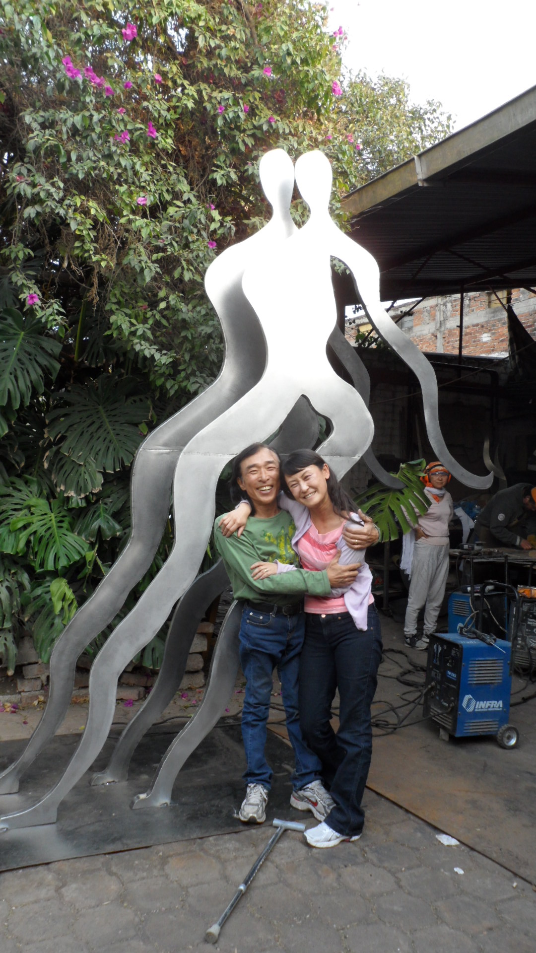 A photo of Won Lee and his wife, Hyon Chu Lee, standing in front of a large-scale artwork titled "Meditators." The piece, which is 80 inches tall, is on permanent display at Vermont State University.
