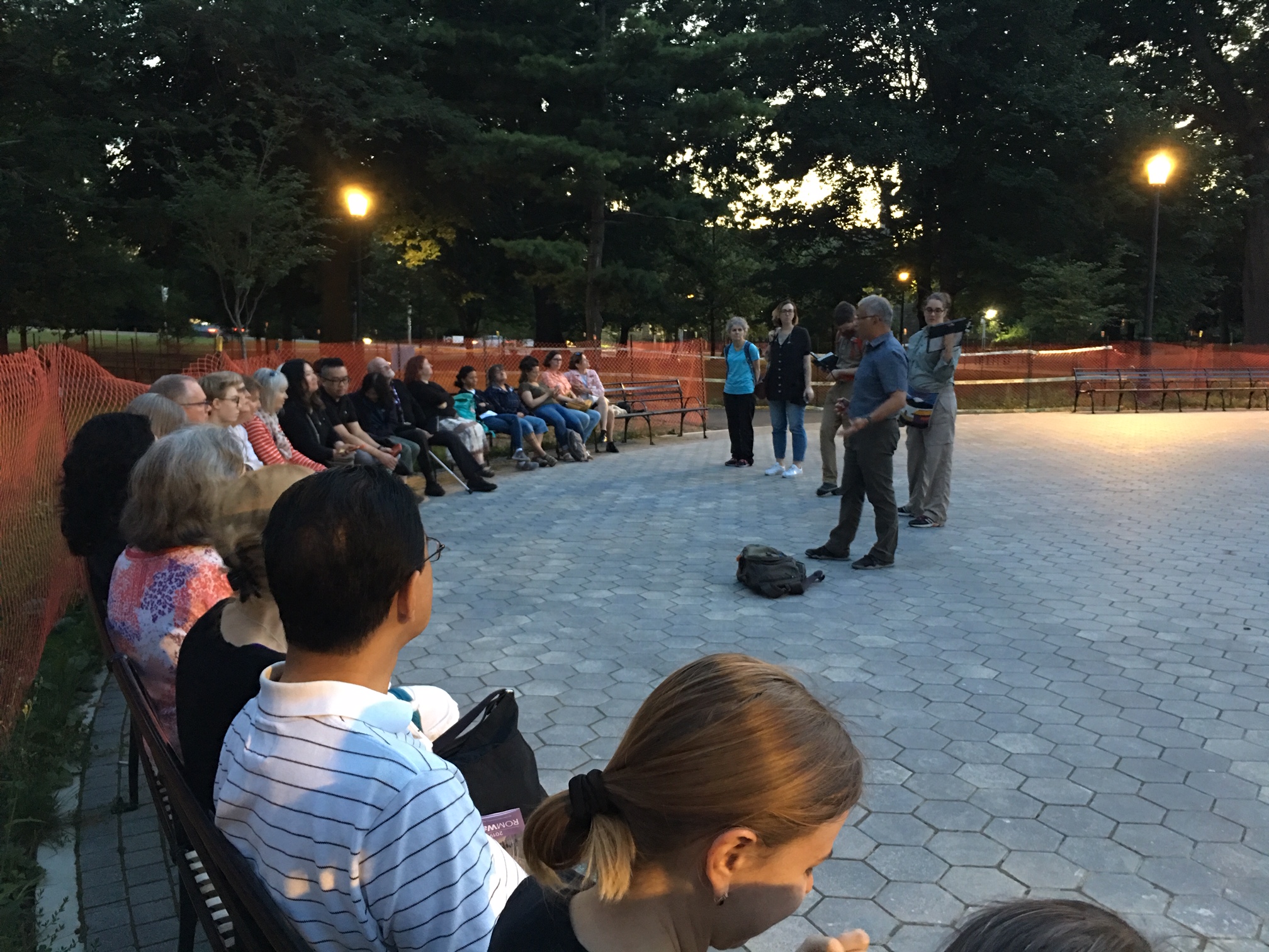 Group of people seated in a park at dusk, listening to a speaker.