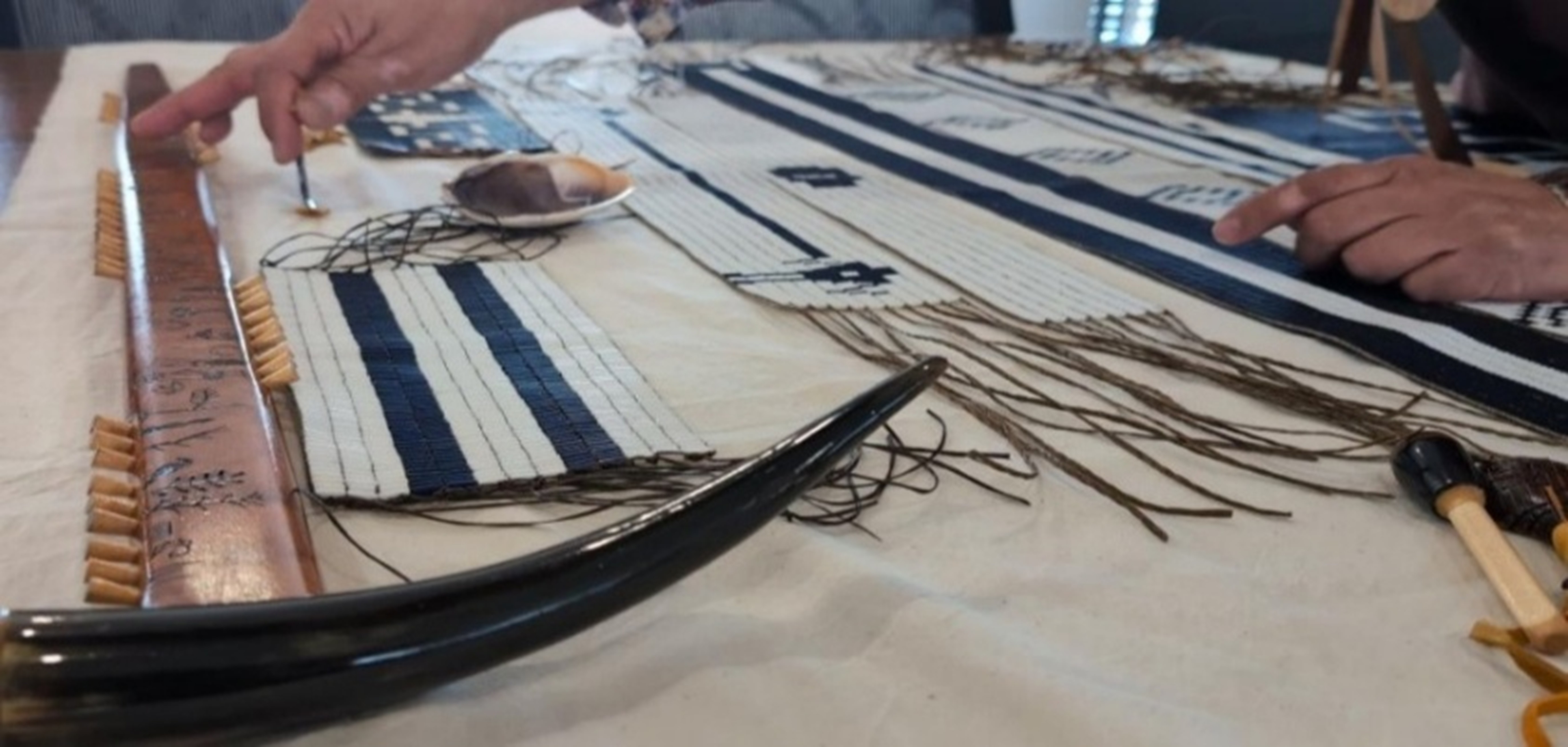 Close-up of hands working on a traditional wampum belt weaving with beads, threads, and tools laid out on a table.