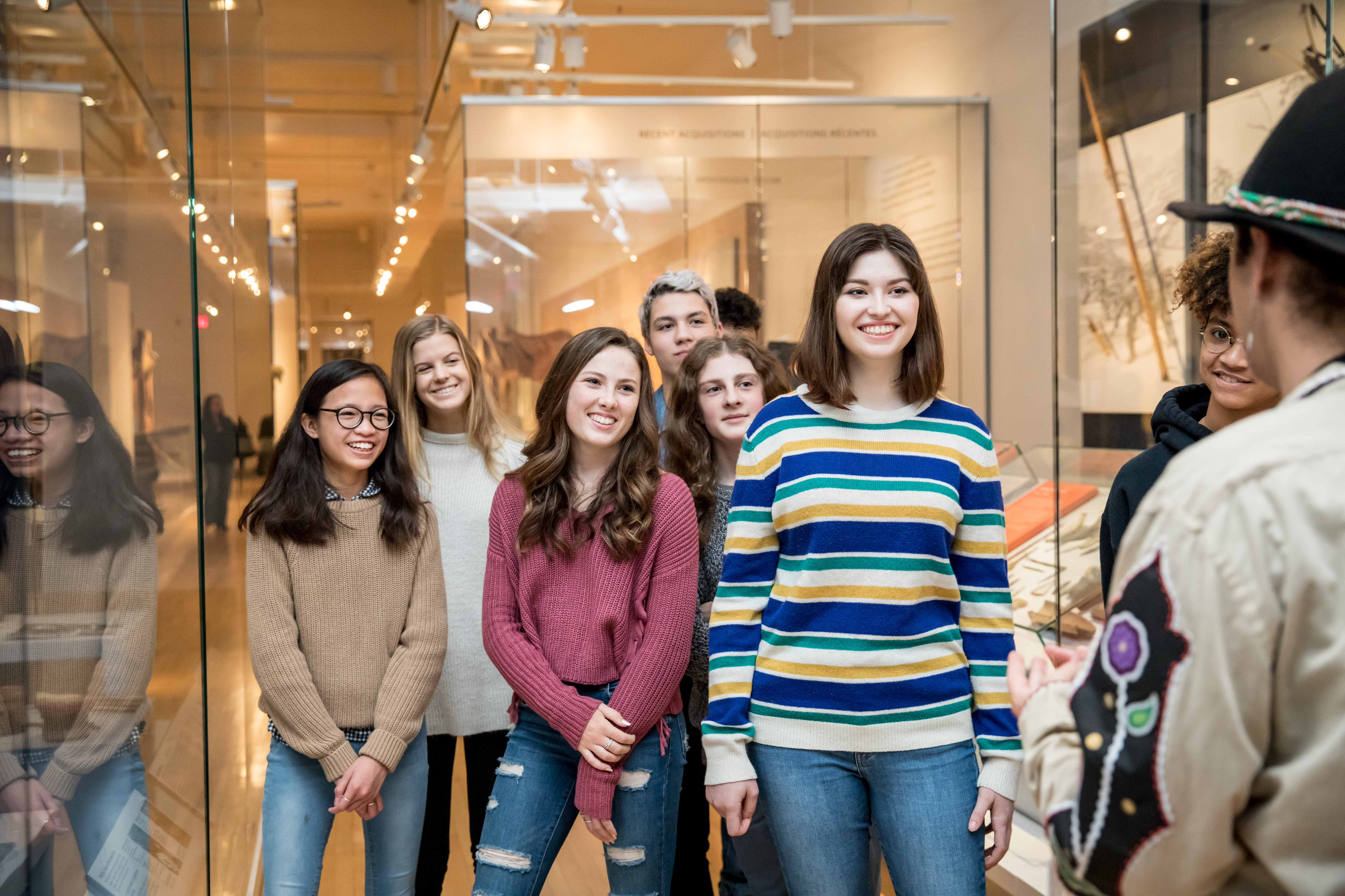 A diverse group of teenage students stands between cases in the First Peoples gallery and smile as they listen to an Indigenous Educator
