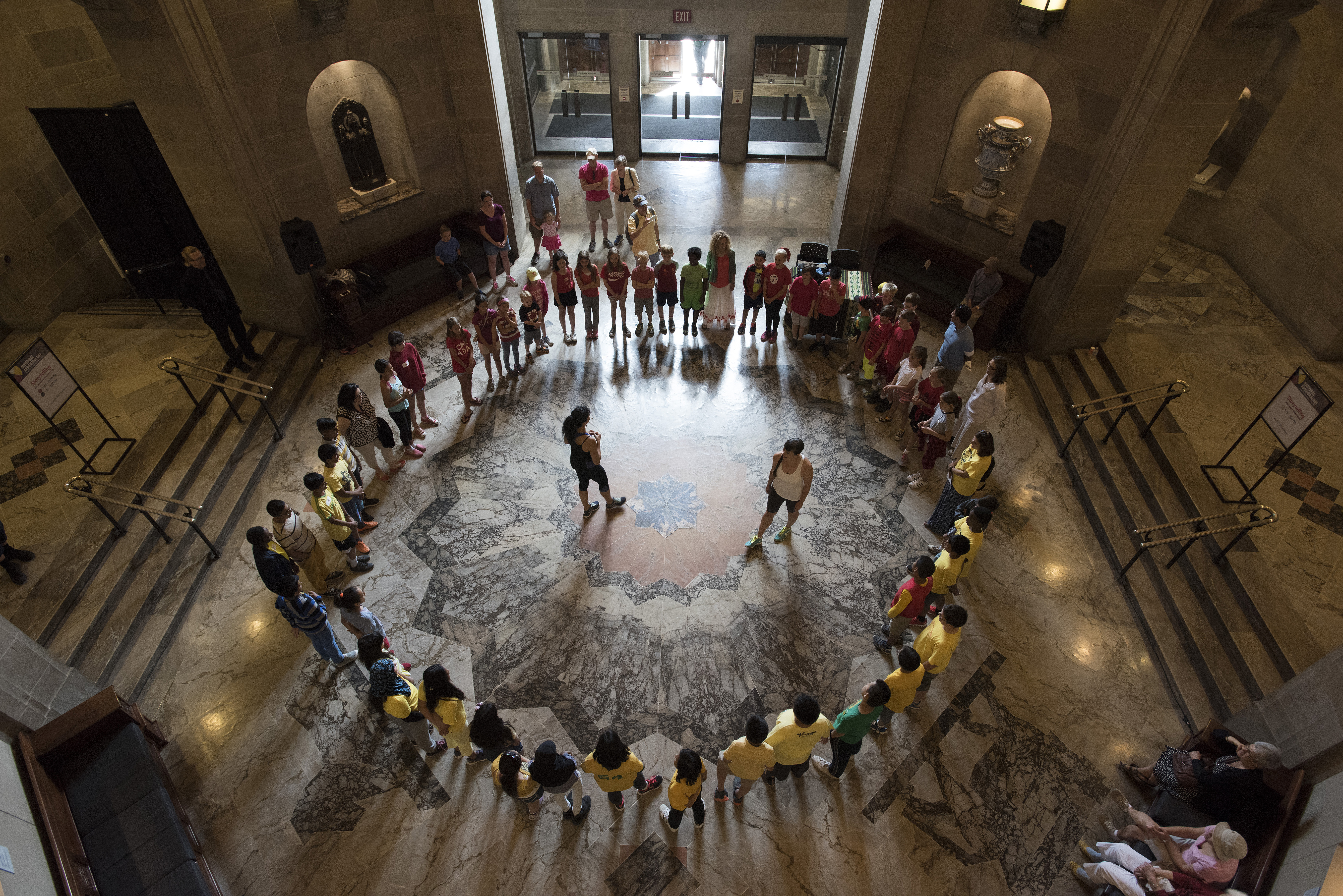 The camera looks down from above at group of students sitting in a circle at the bottom of a round ornate room