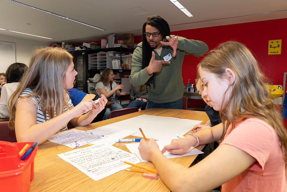 Two students sitting at a table work on a plan for a porcupine quill stitching game. Another student looks up at an adult making a square shape with his fingers.