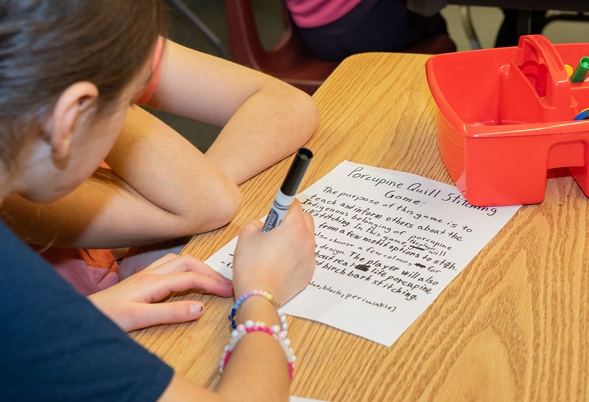 Students sitting at a table write a plan for a porcupine quill stitching game