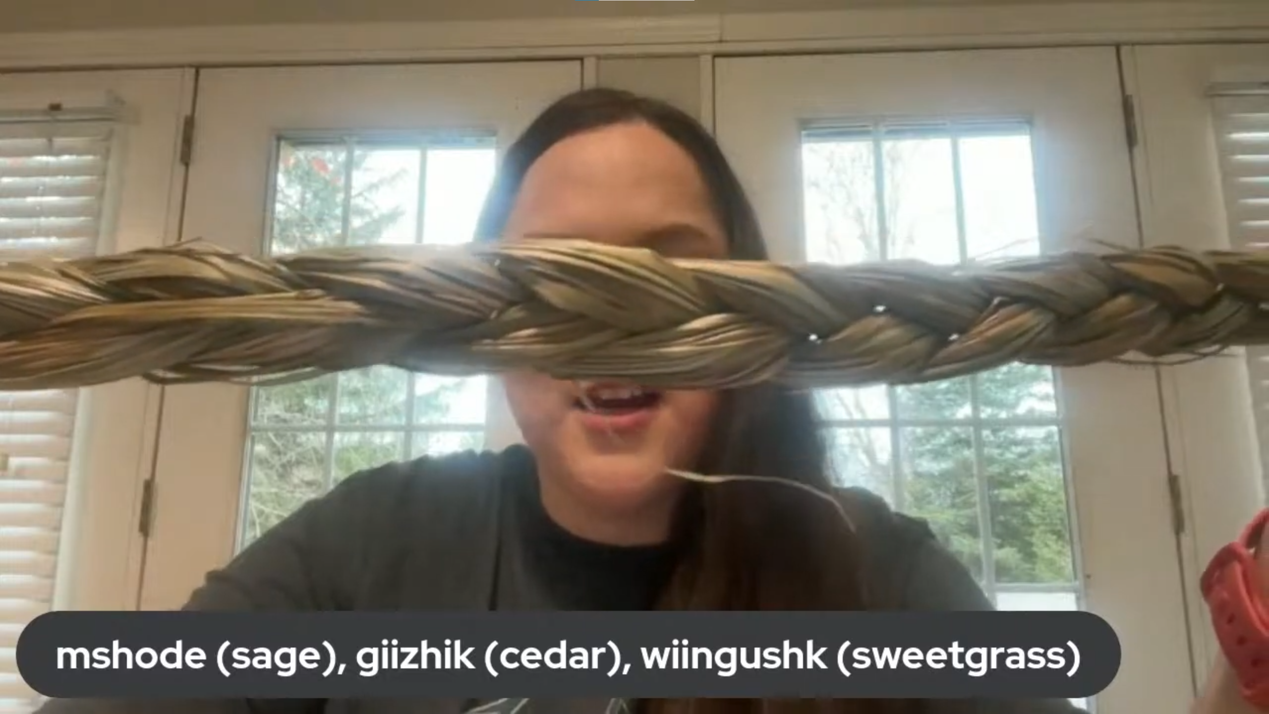 An Indigenous woman holds up braided sweetgrass. A caption on the bottom of the screen reads mshode (sage{, giishik (cedar), wiingushk (sweetgrass)