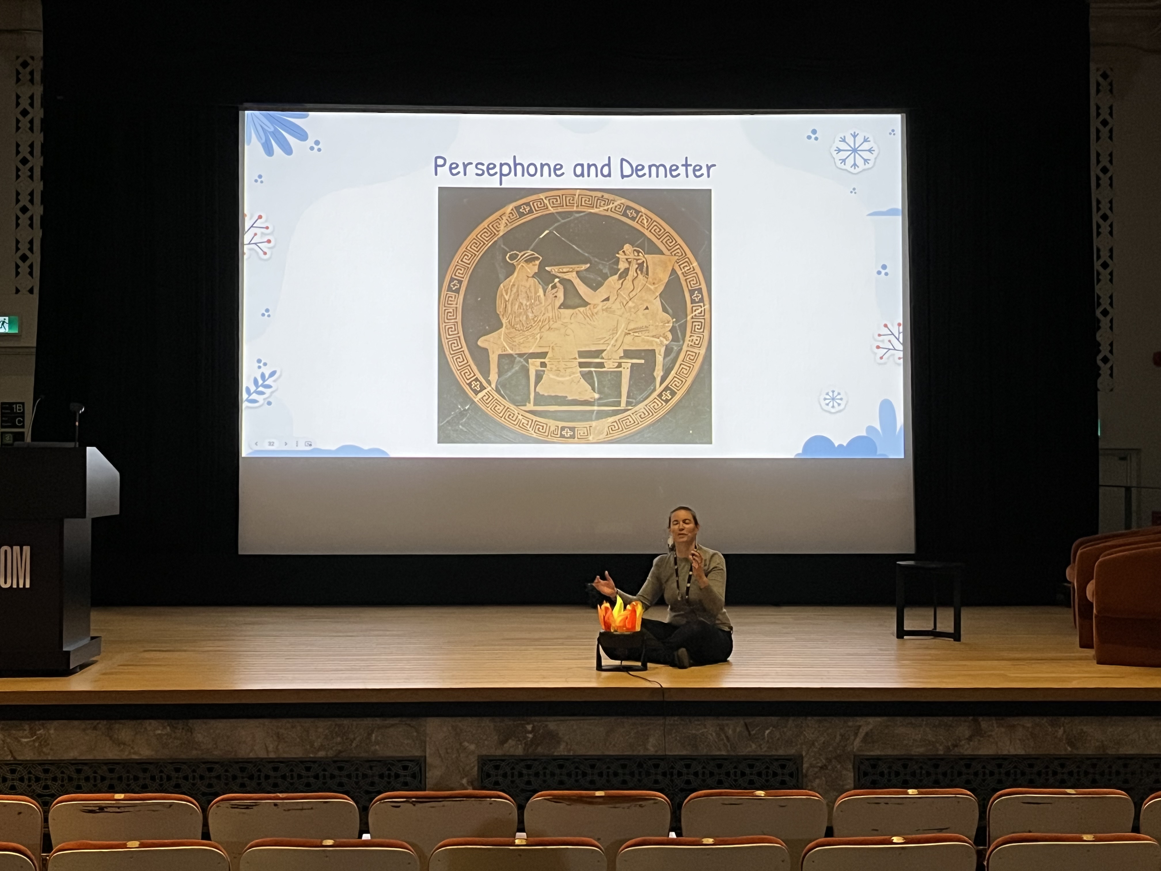 A woman sits on a theatre stage next to an artificial campfire. A screen behind her shows a slide titled Persephone and Demeter, with an image of two goddesses depicted in red-figure Greek pottery.