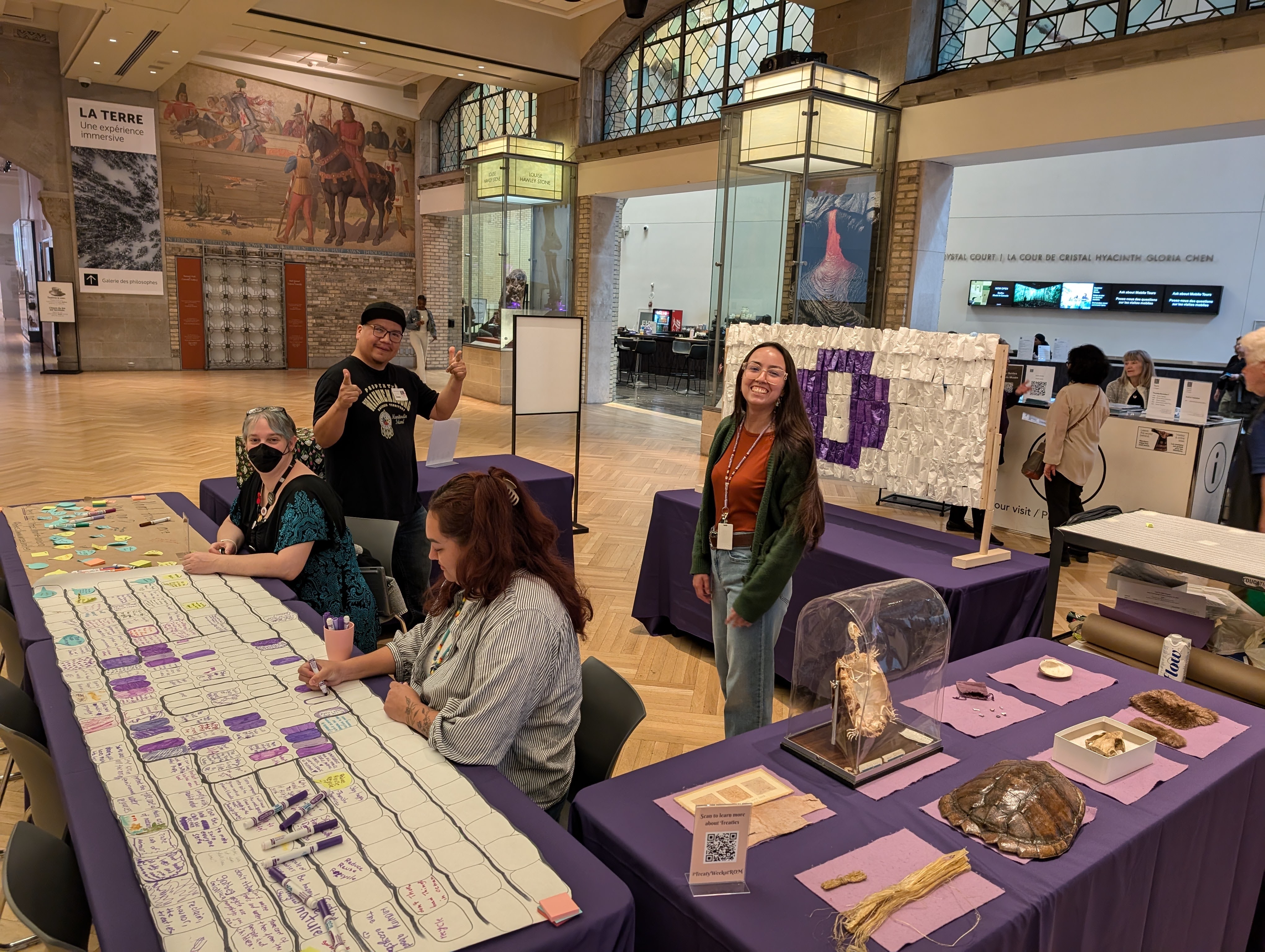 Activation Station A station in a large gallery covered in activities, objects, and a white wampum belt with a purple circle at the back. Staff man the activities.