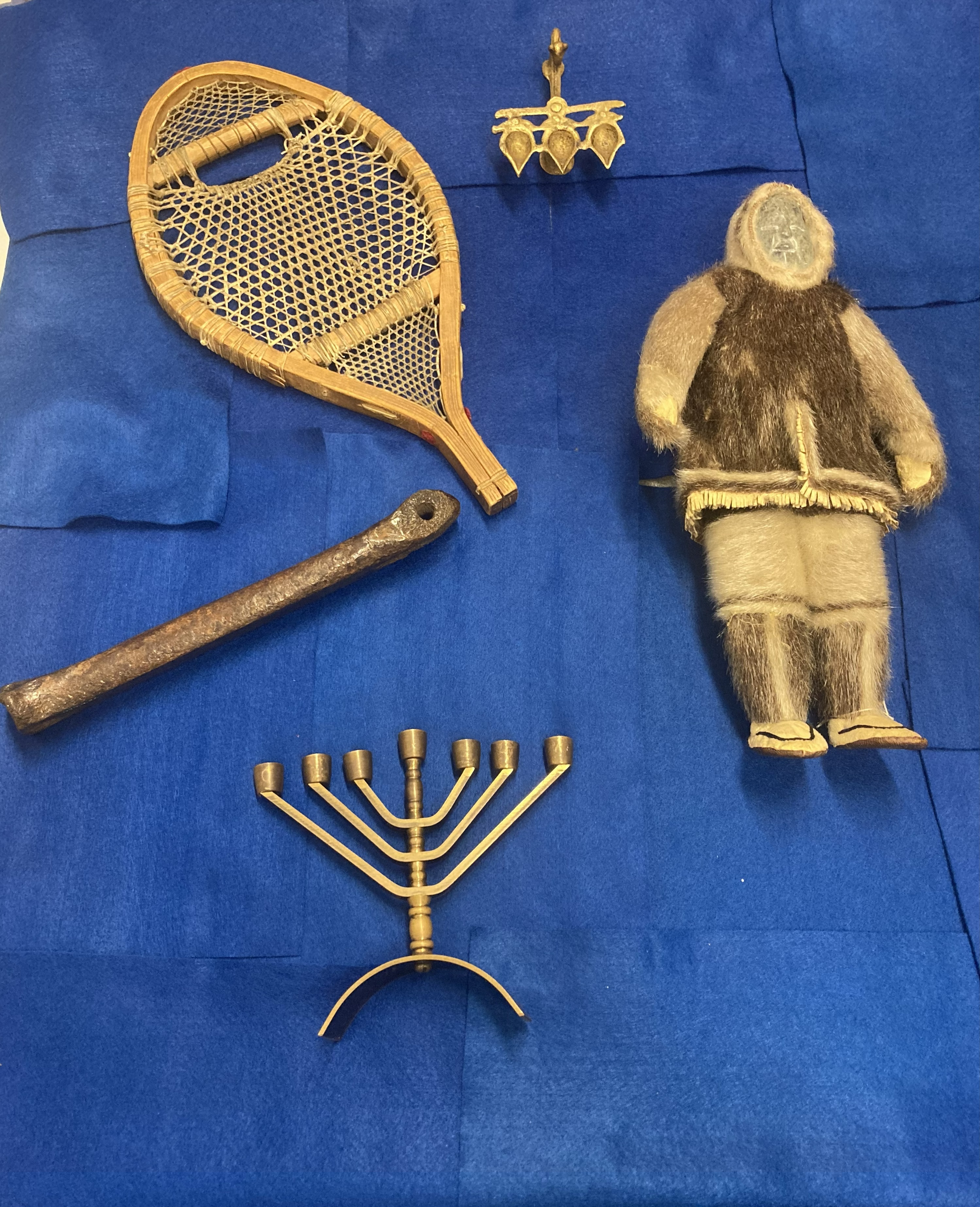 A selection of objects on a blue background.  The objects include an Inuit stone and sealskin doll, a Hannukiah, a medieval bone skate, a snowshoe, and a diya lamp.