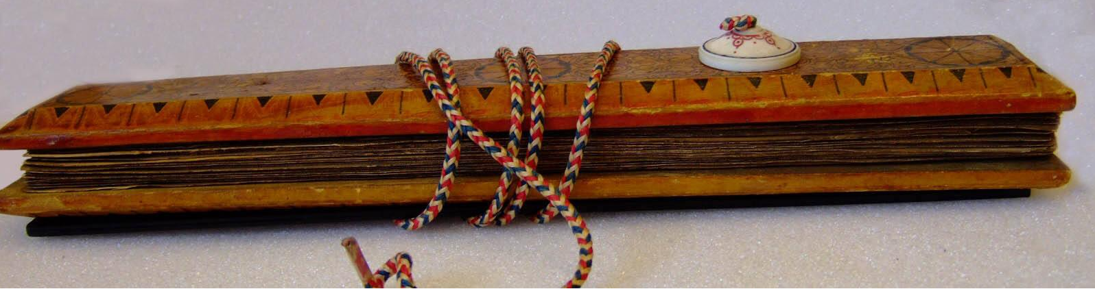 A side view of a manuscript sandwiched between two wooden cases. Decorations visible on the top case include black circles, and alternating lines and triangles. A white button on the upper case secures a red, white, and blue cord that wraps around both cases and holds the manuscript together.