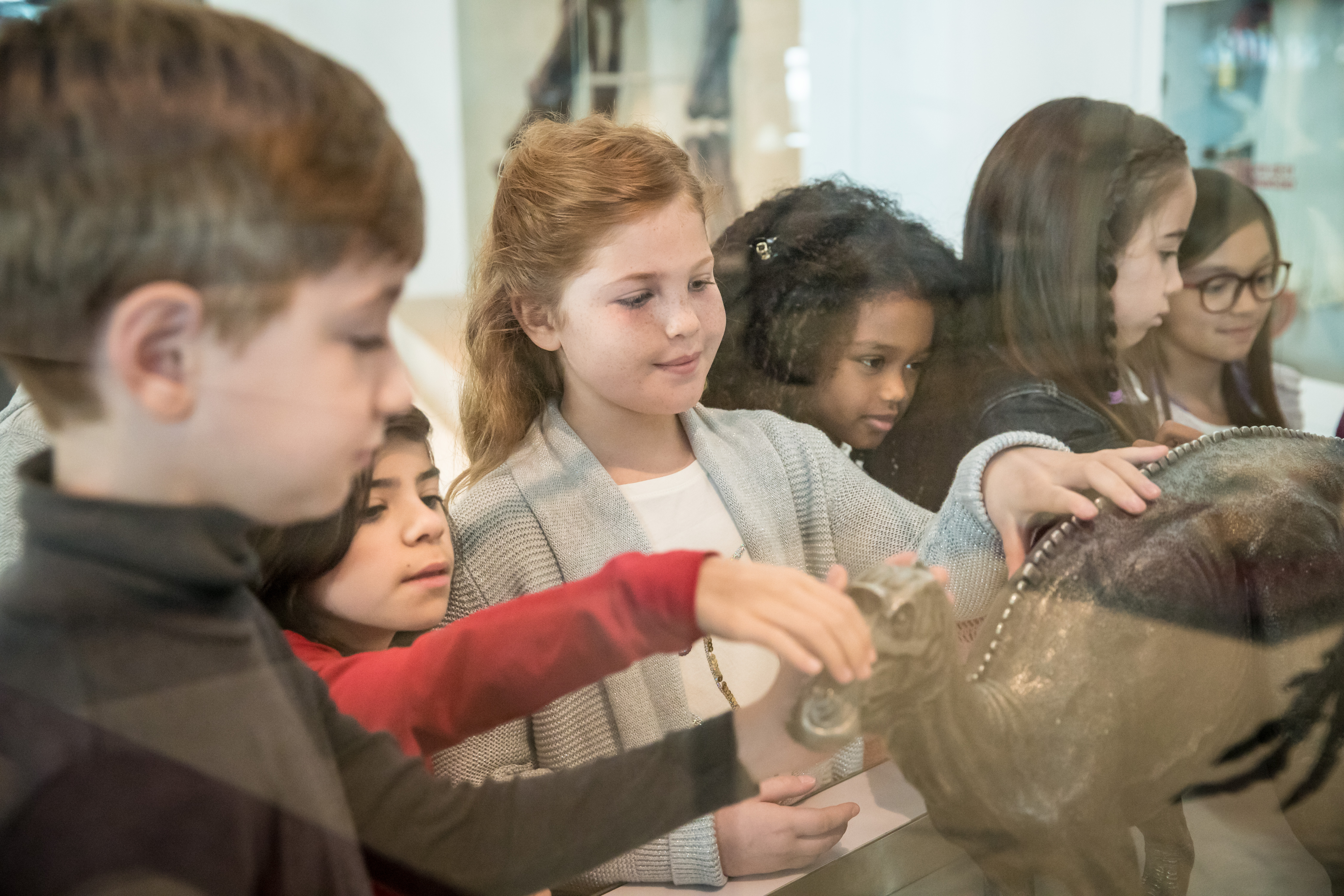 Several young students in a diverse class touching a bronze statue of a maiasaur.