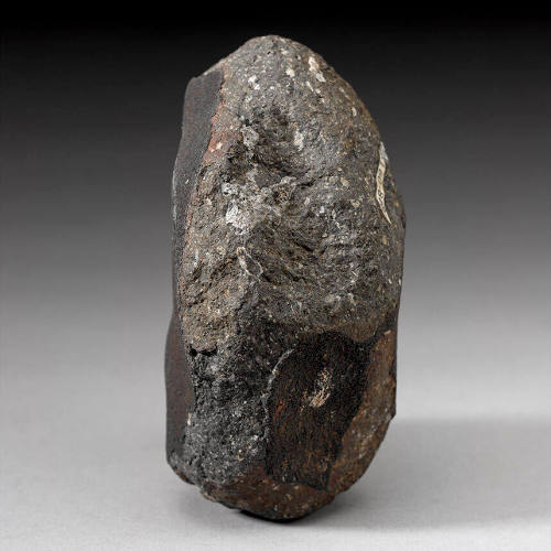 A rock that is taller than it is wide. It is dark grey with white specks, and some parts have a black crust on them.