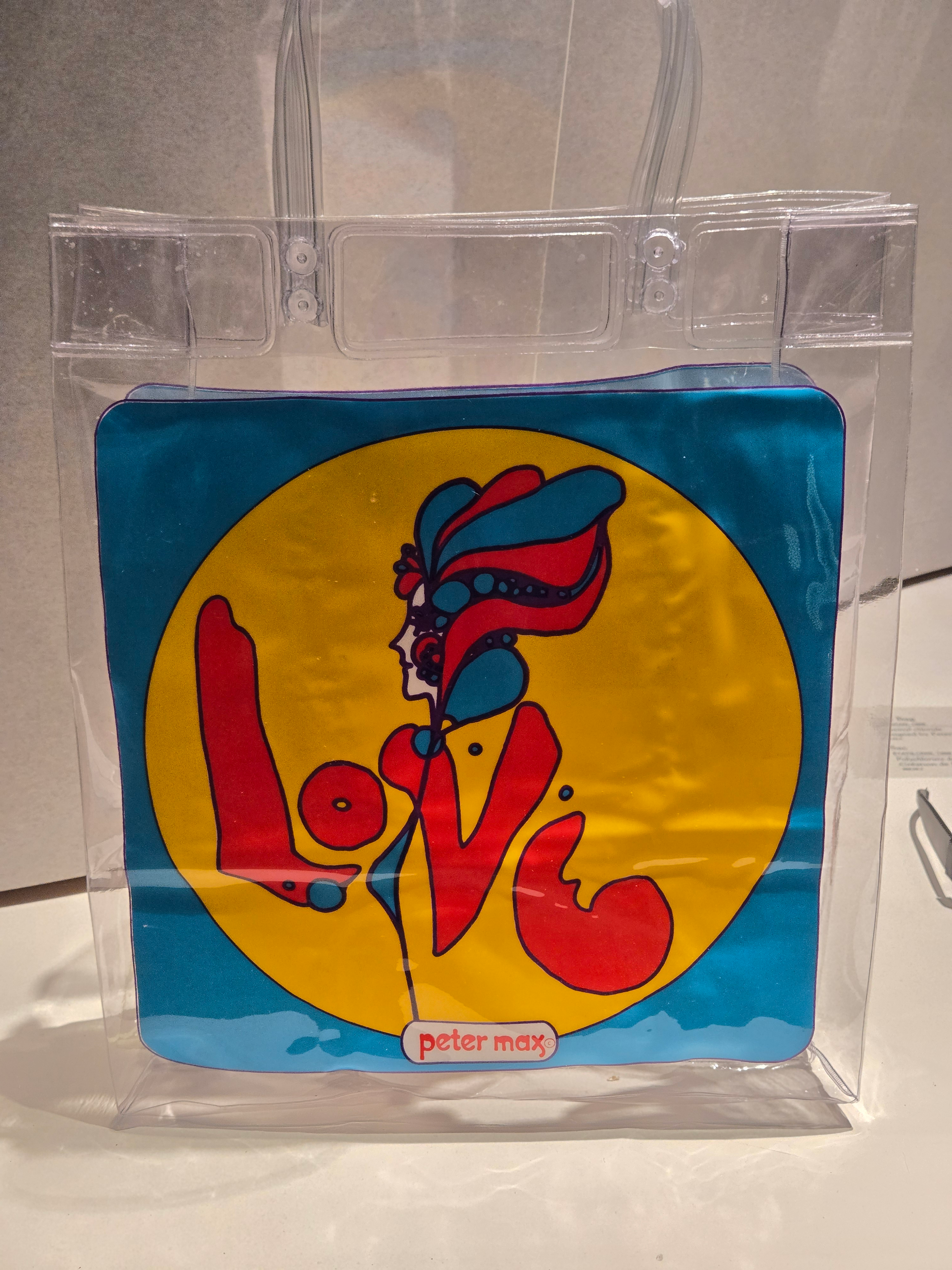 A clear plastic bag in a museum case. On the front of the bag is an image of a woman in a yellow circle against a blue background. The woman’s arms and coat also spell the word LOVE. Peter max is printed at the bottom.