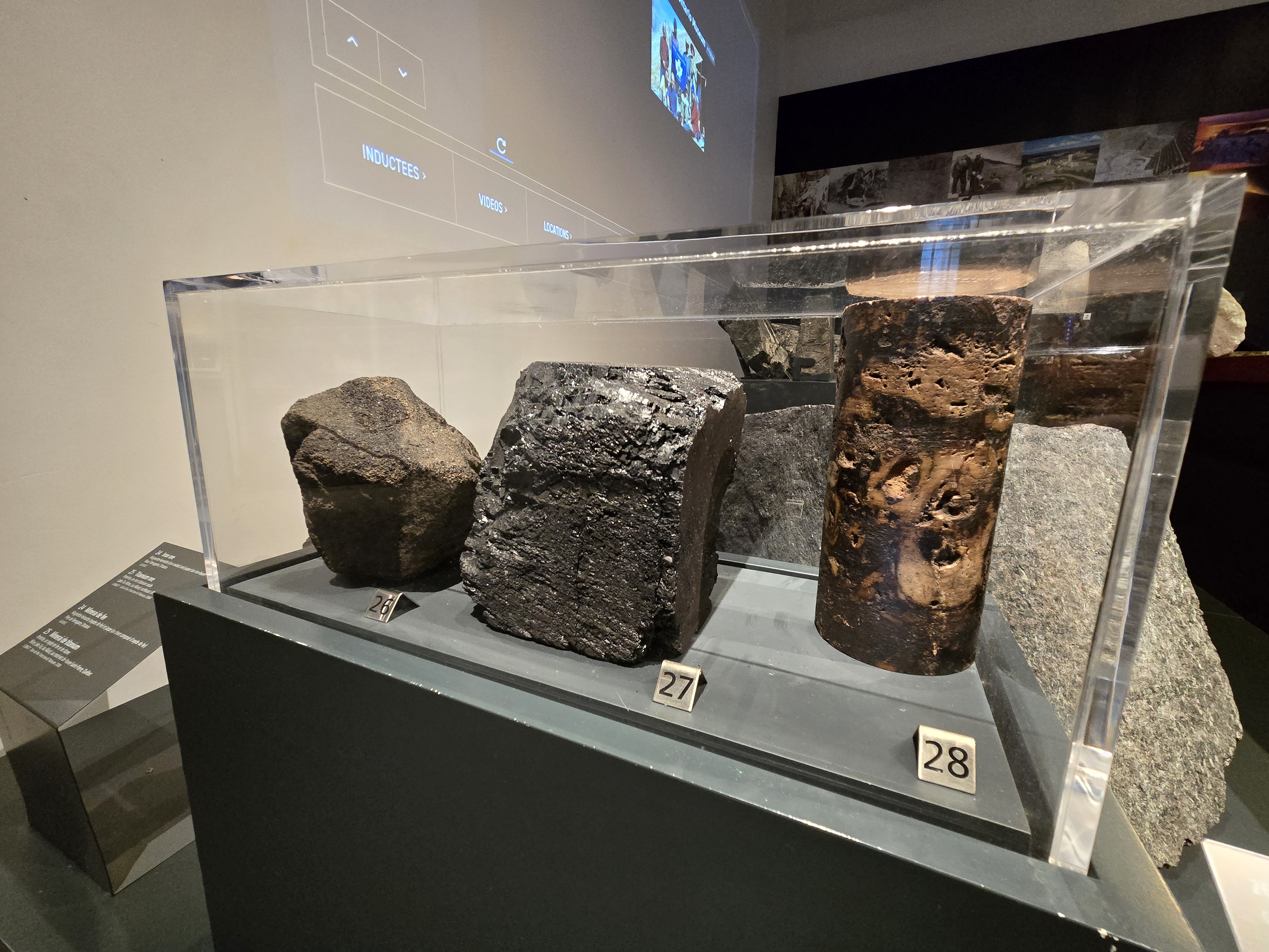 A case in a museum gallery holds three rocks. One is dull brown/black and rounded, one is black and shiny, and one has brown tones with lighter patches against darker rock.