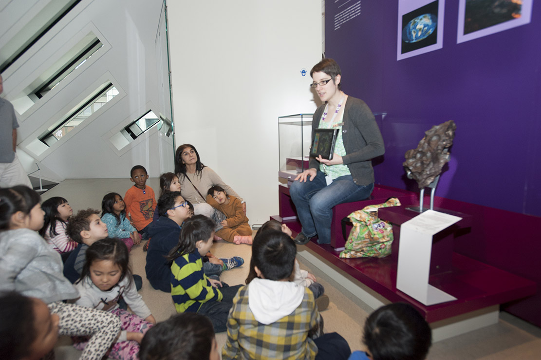 Several young students in a diverse class seated in front of an Educator who is showing them images on a tablet.