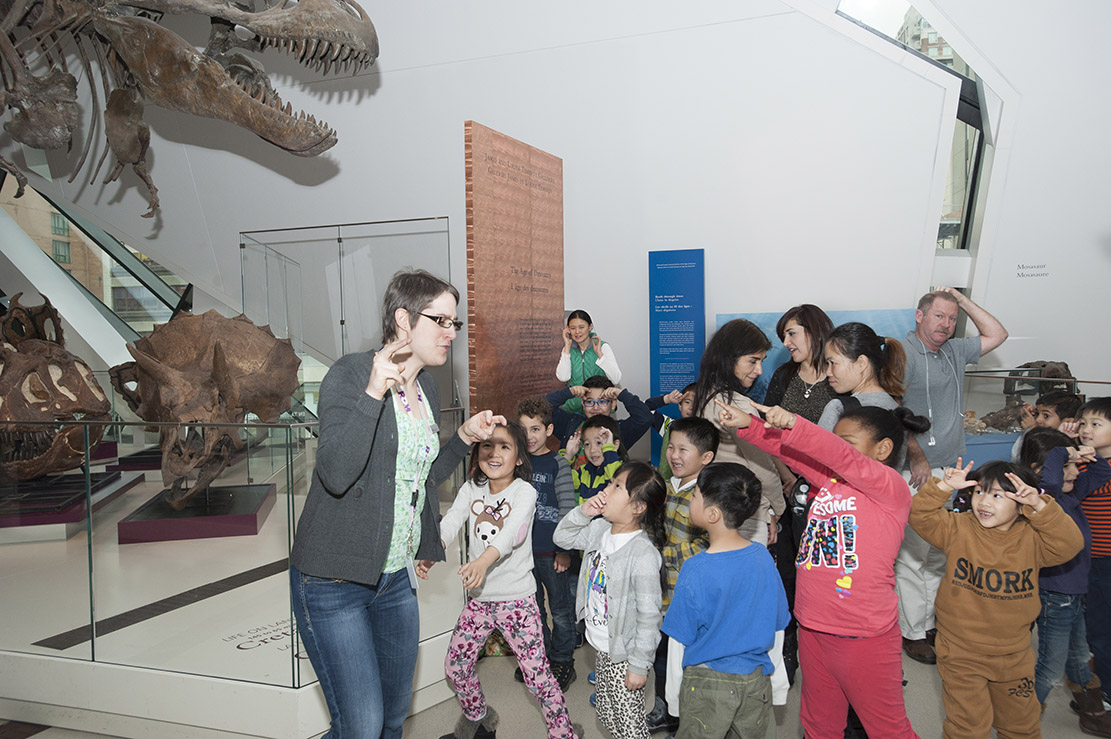 Several young students in a diverse class using fingers to make horns on their foreheads and pointing at teacher who is making claws with her fingers.