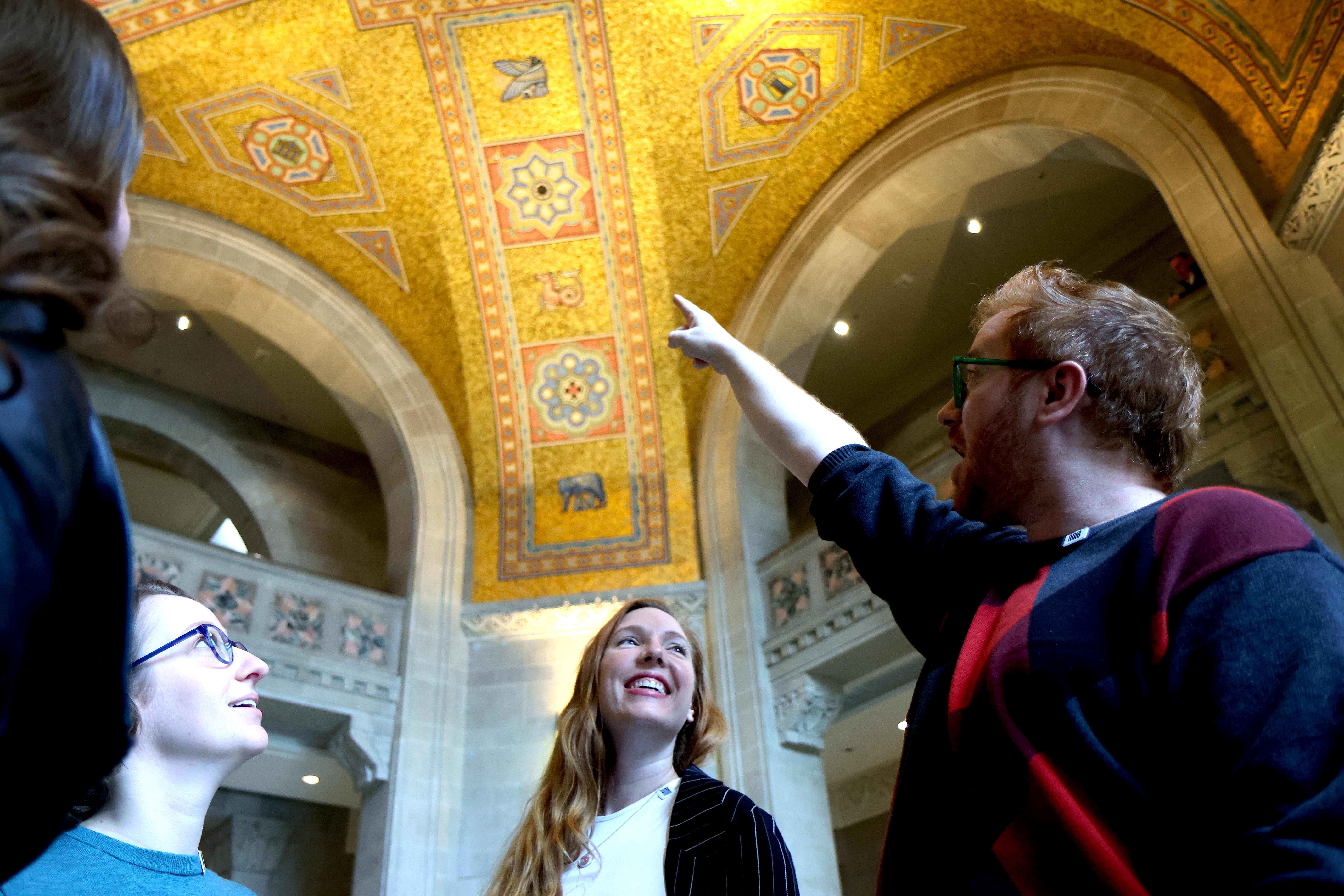 Three happy people look up at an ornate golden ceiling as one person points out a detail.