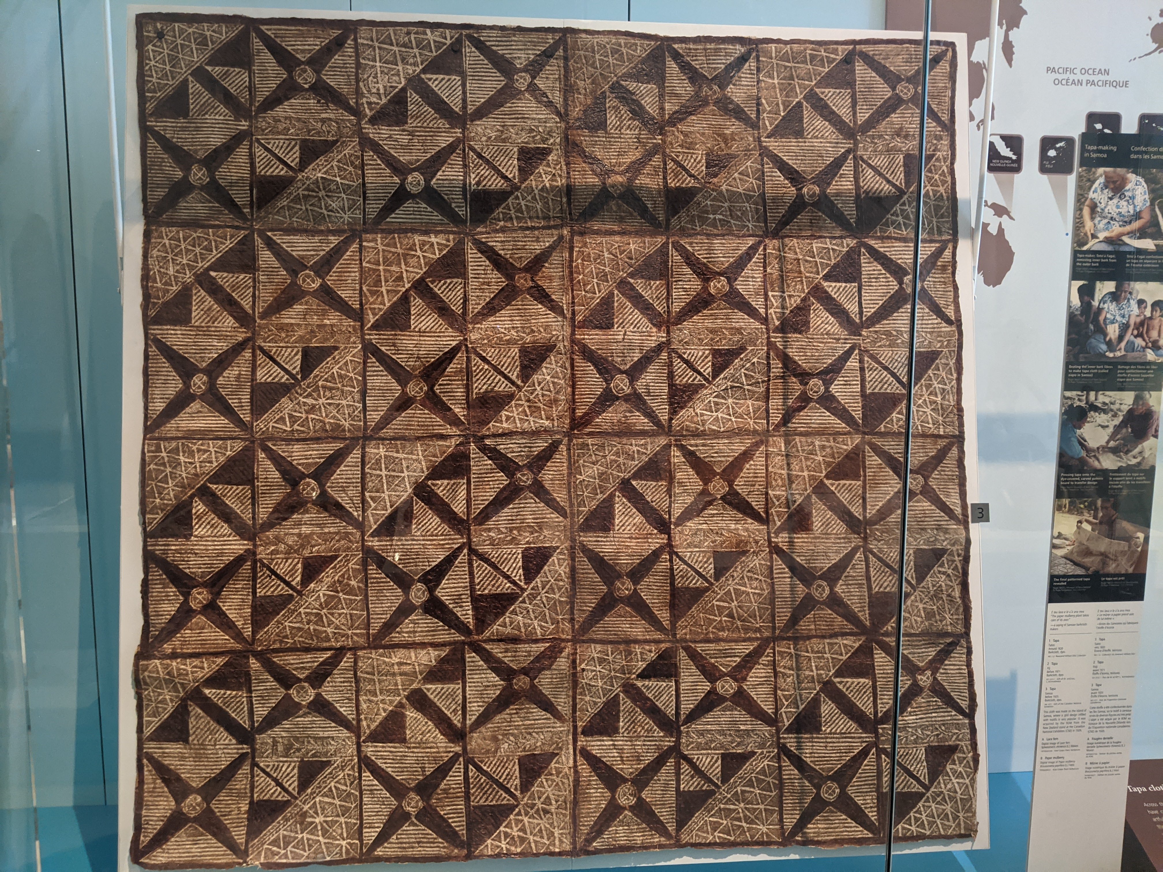 A large square of brown and beige cloth hangs in a museum display case. The cloth has intricate geometric patterns made up of lines, squares, and triangles.