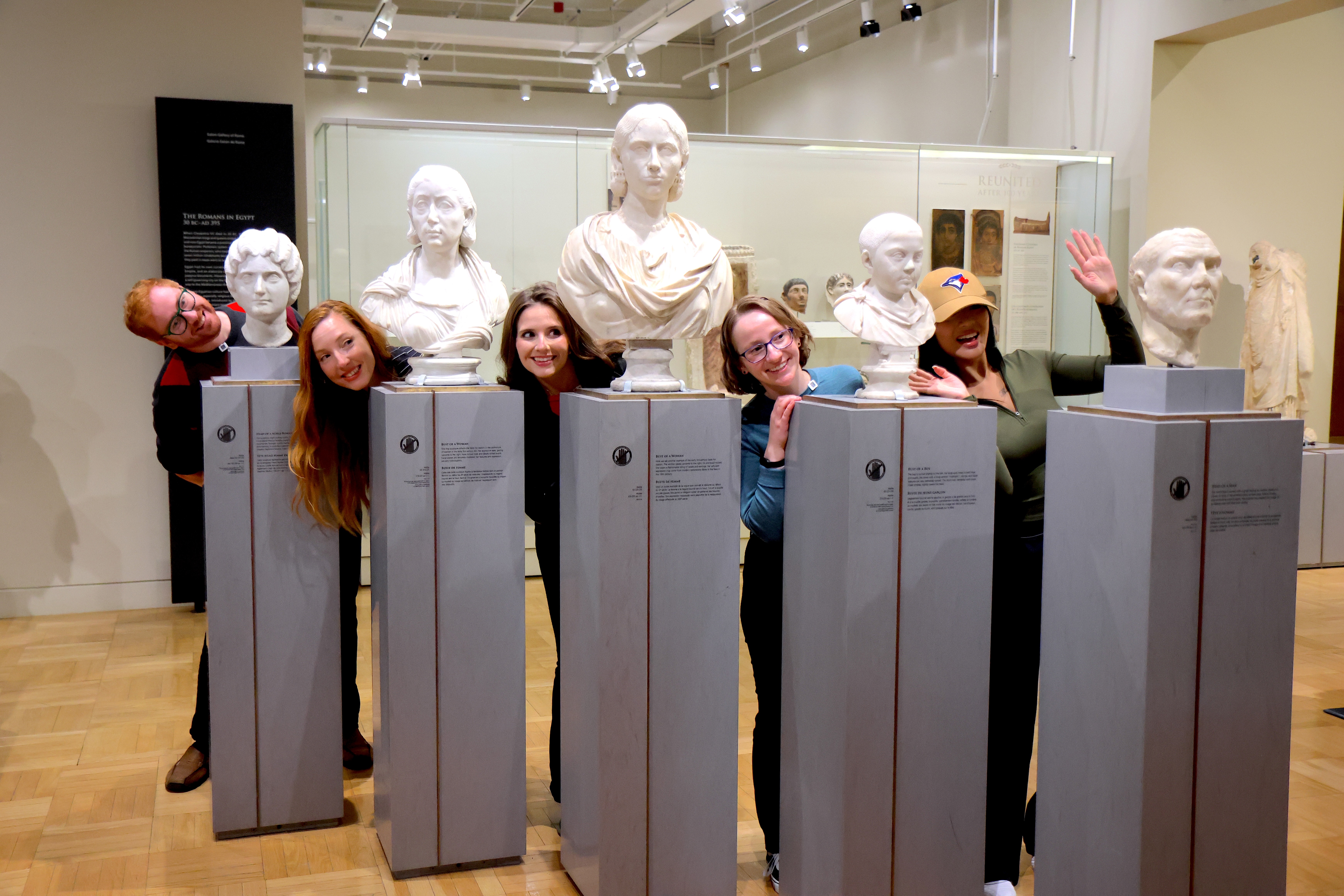 "Un groupe de personnes pose de manière ludique en se penchant entre des bustes de marbre classiques dans une galerie de musée.