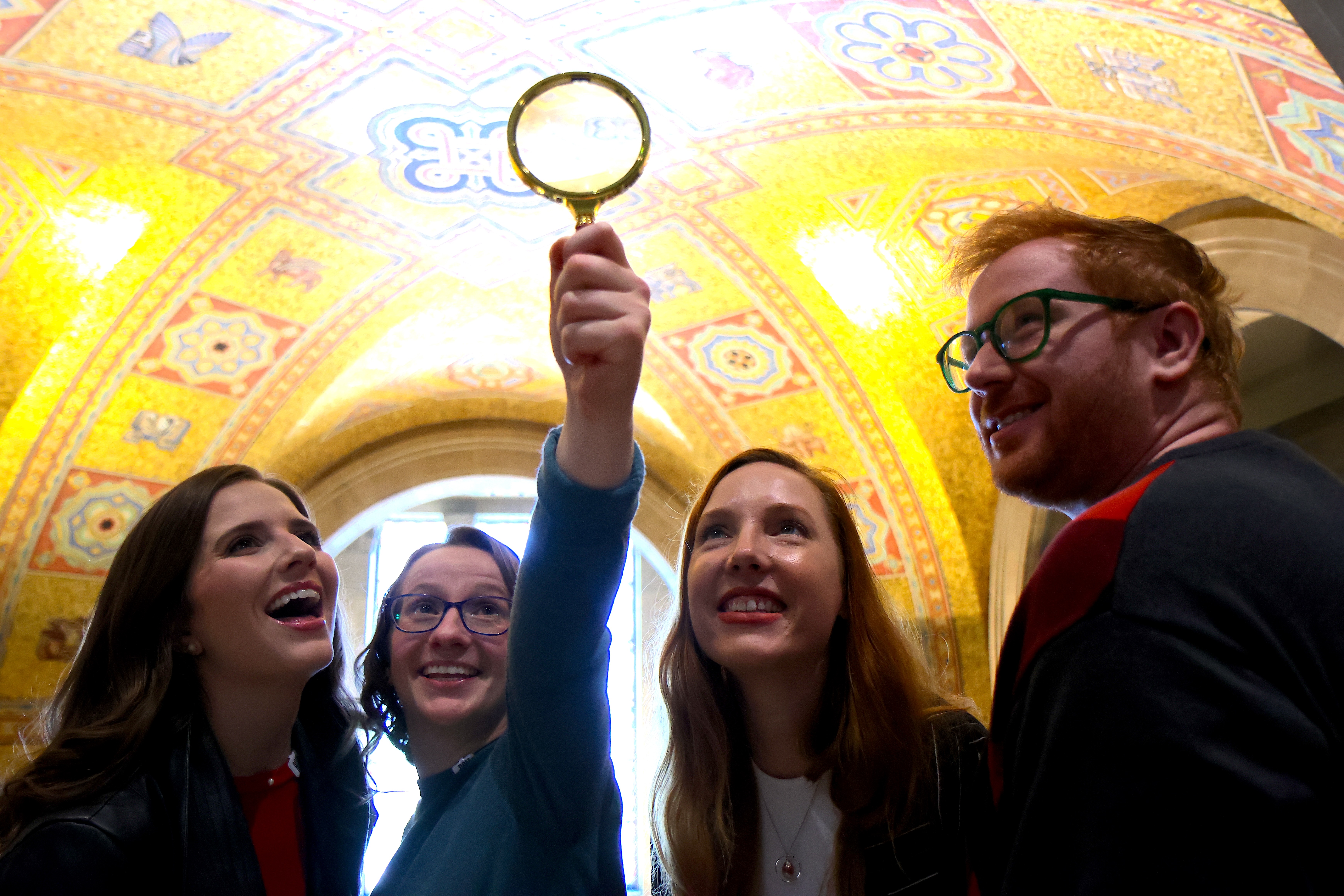 Un groupe de personnes sourit et brandit une loupe en explorant le plafond doré et orné de la rotonde du musée.
