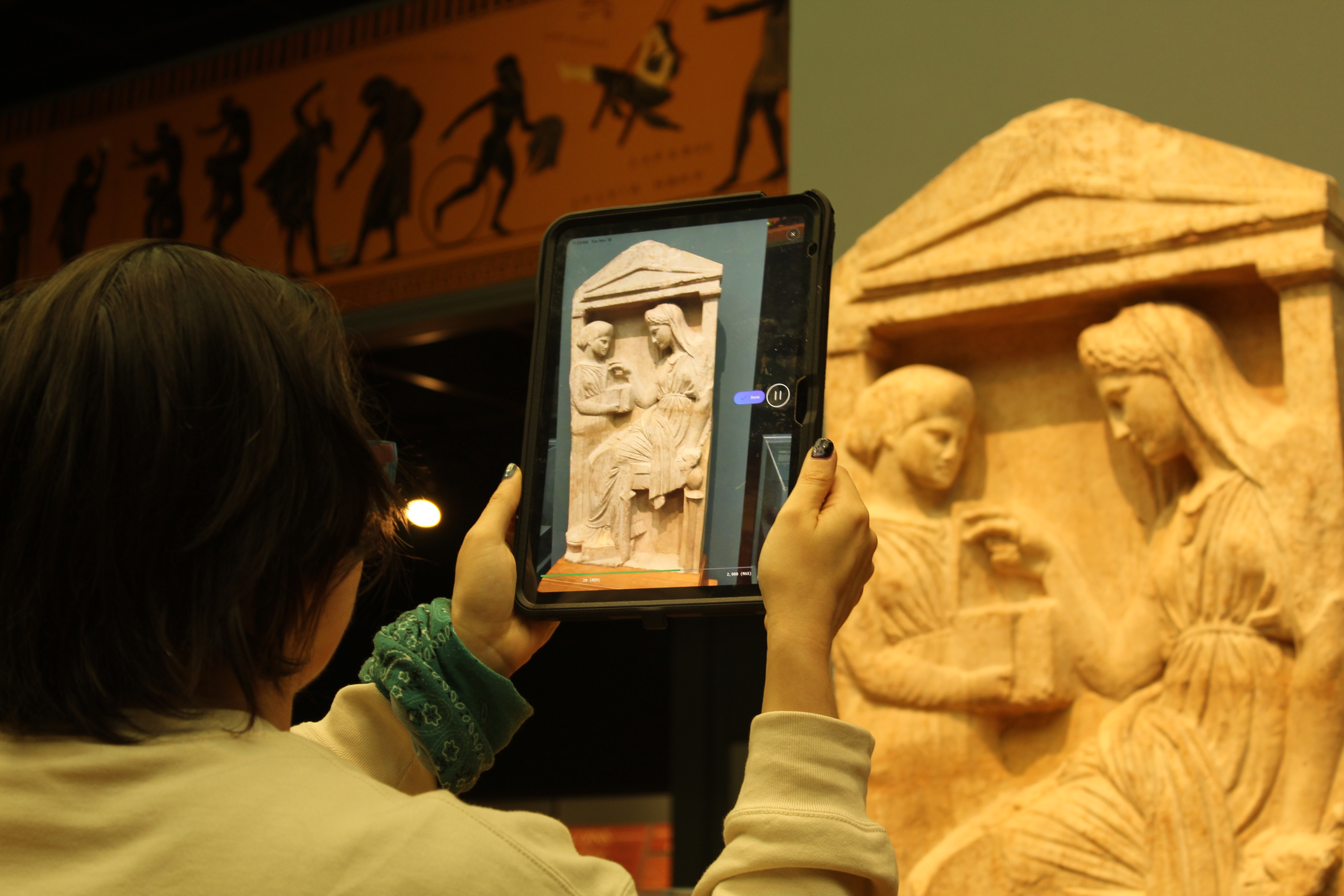 A person holds a tablet up to scan a Greek carving of a woman choosing jewellery from a box.