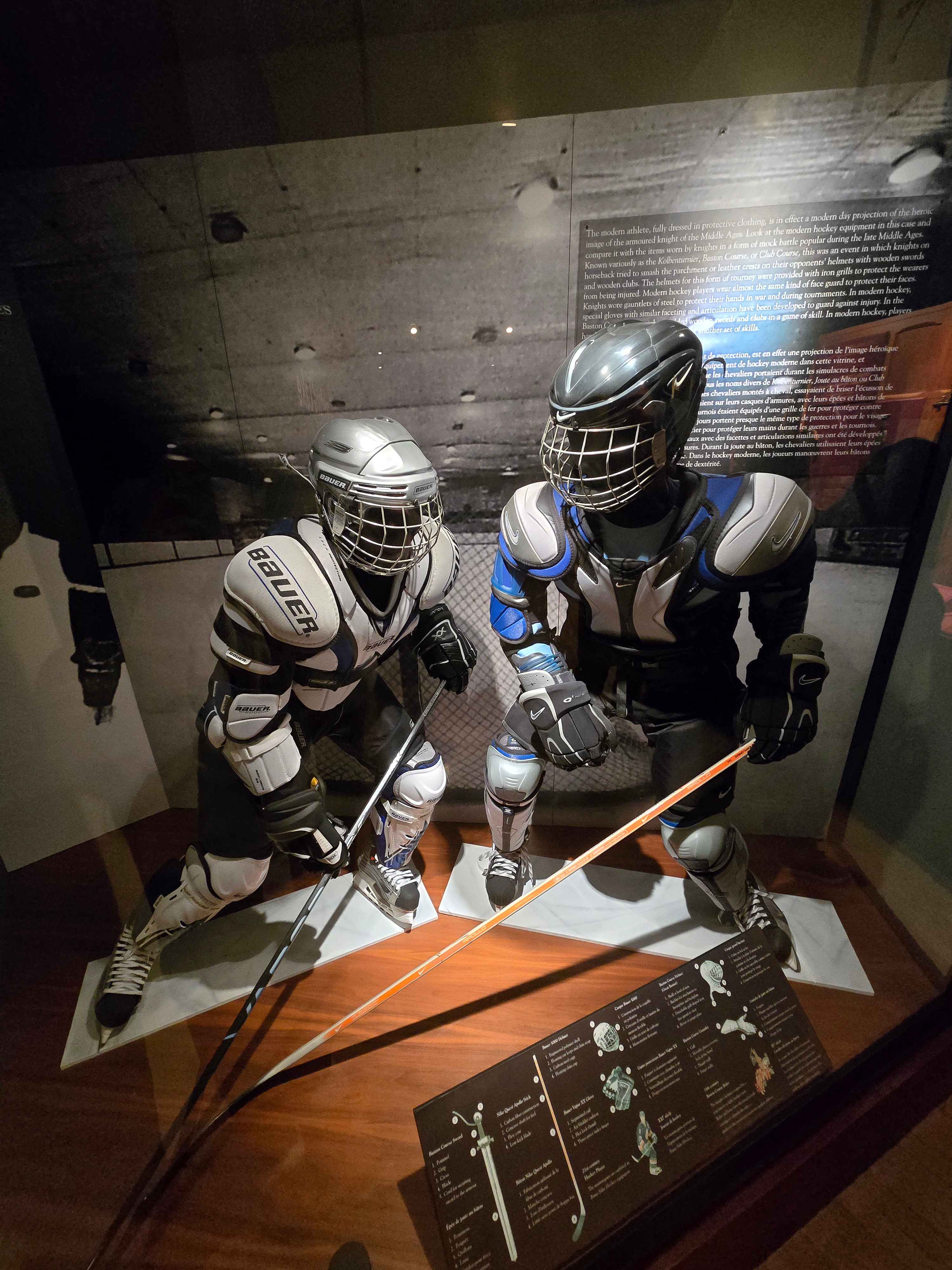 Two mannequins dressed in Bauer hockey gear, including shoulder pads, shin pads, gloves, skates, and helmets. Both mannequins hold hockey sticks and are in dynamic action poses.