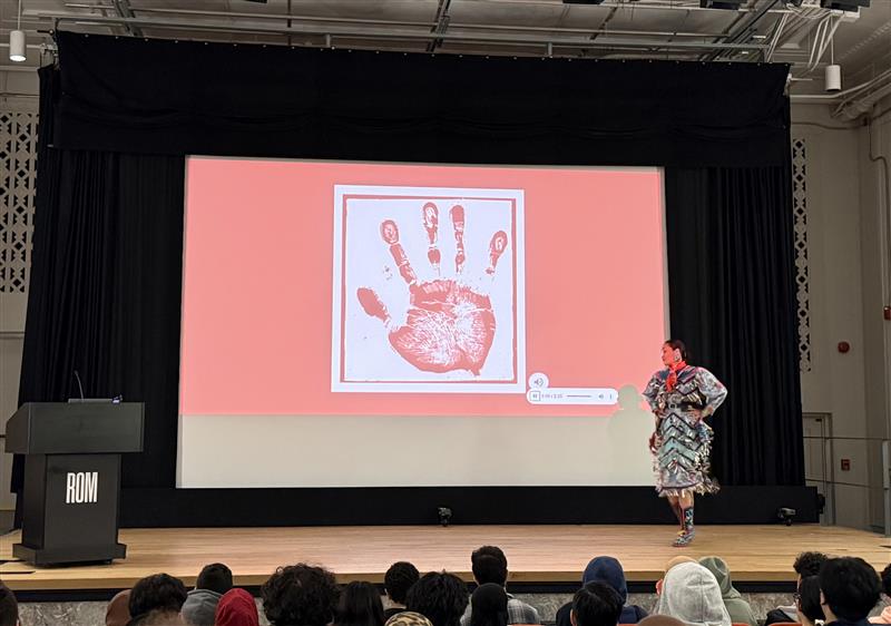 An Indigenous woman wearing a jingle dress stands on a stage in a theatre. The screen behind her shows a white handprint on a red screen.