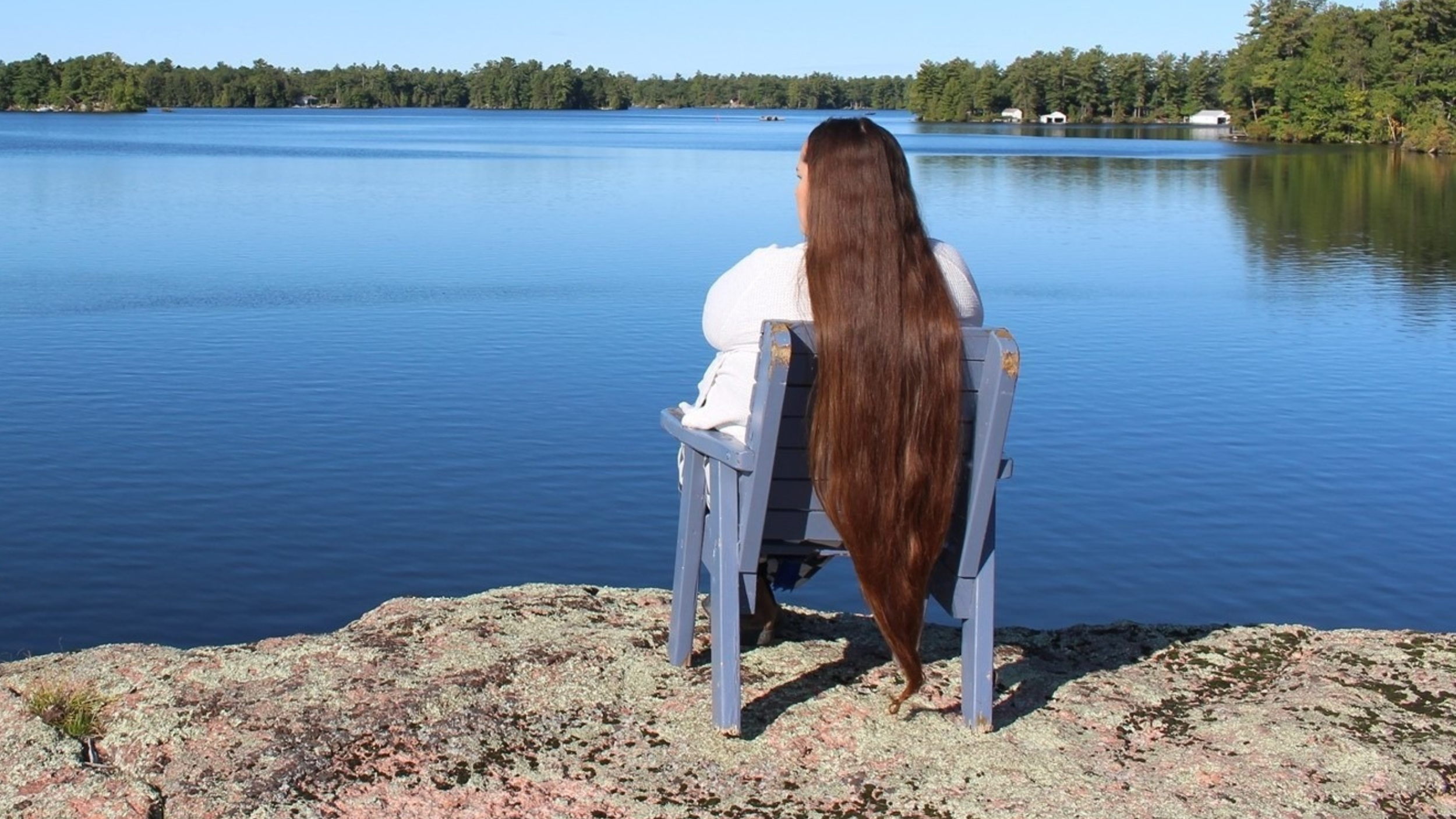 A woman with long hair sits on a chair overlooking a glassy blue lake.