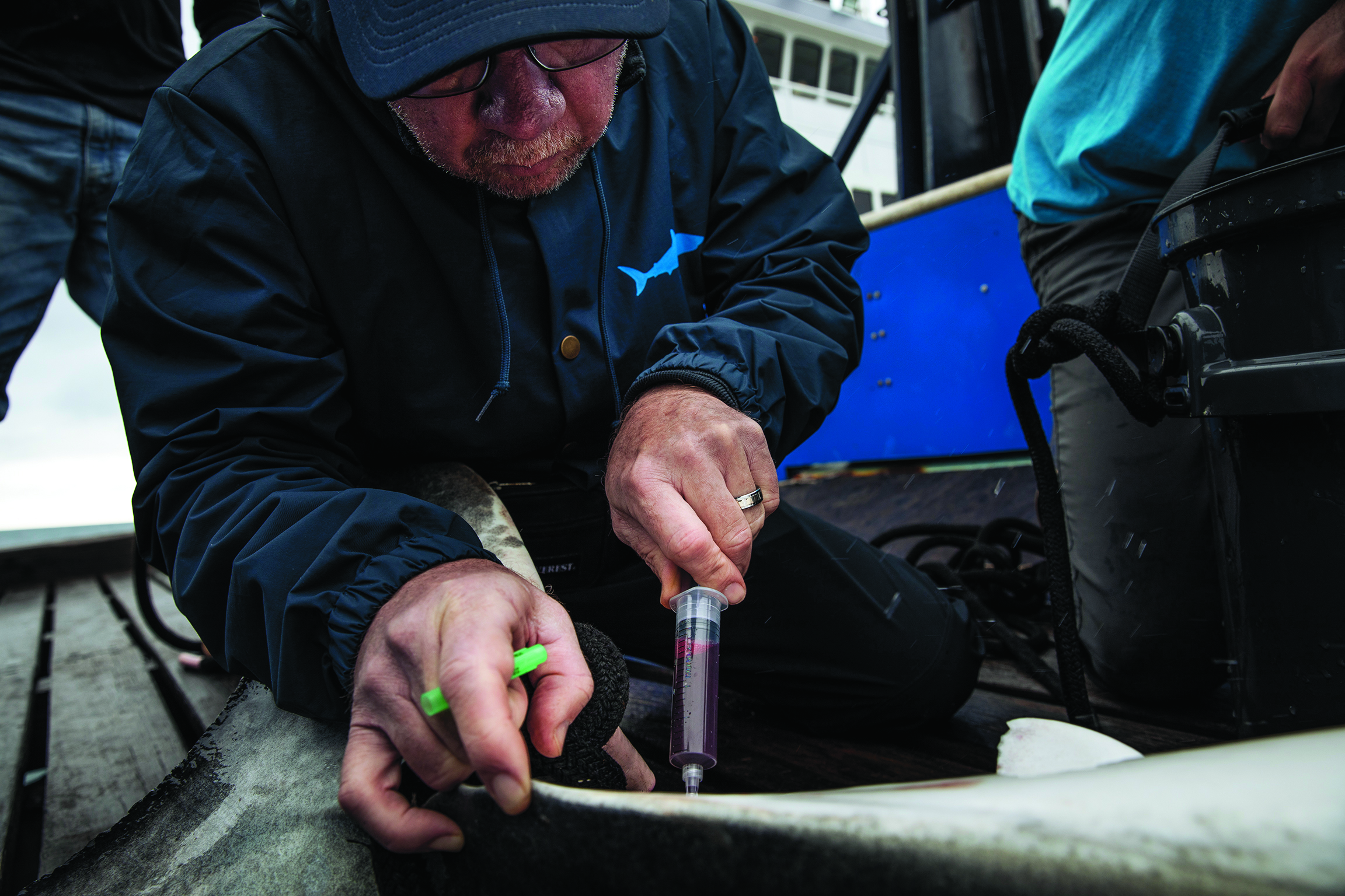 Man wearing glasses and a blue jacket crouched down holding a small container and a syringe.
