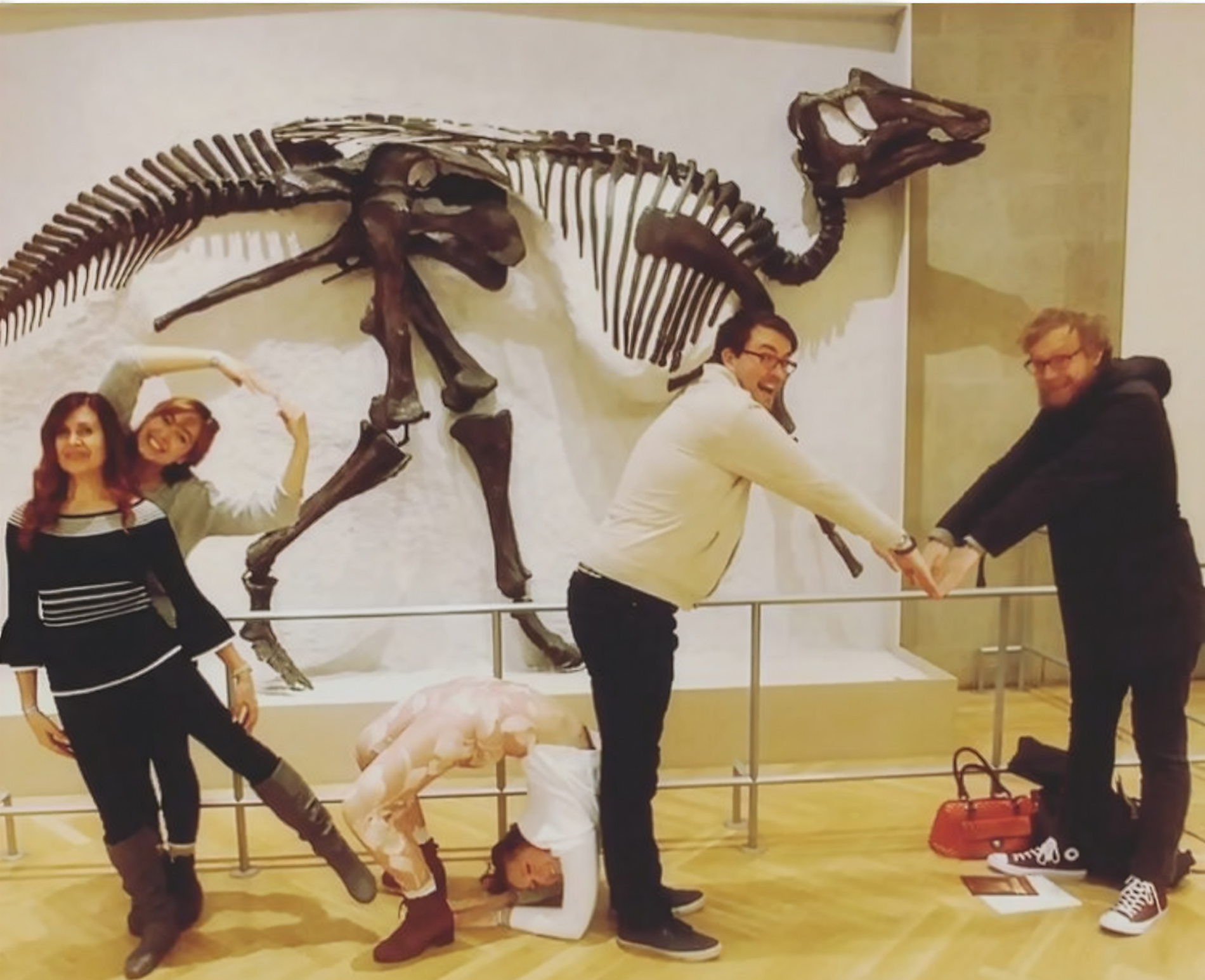 Un groupe de personnes prend des poses ludiques devant un squelette de dinosaure, en arrangeant leur corps pour épeler des formes.