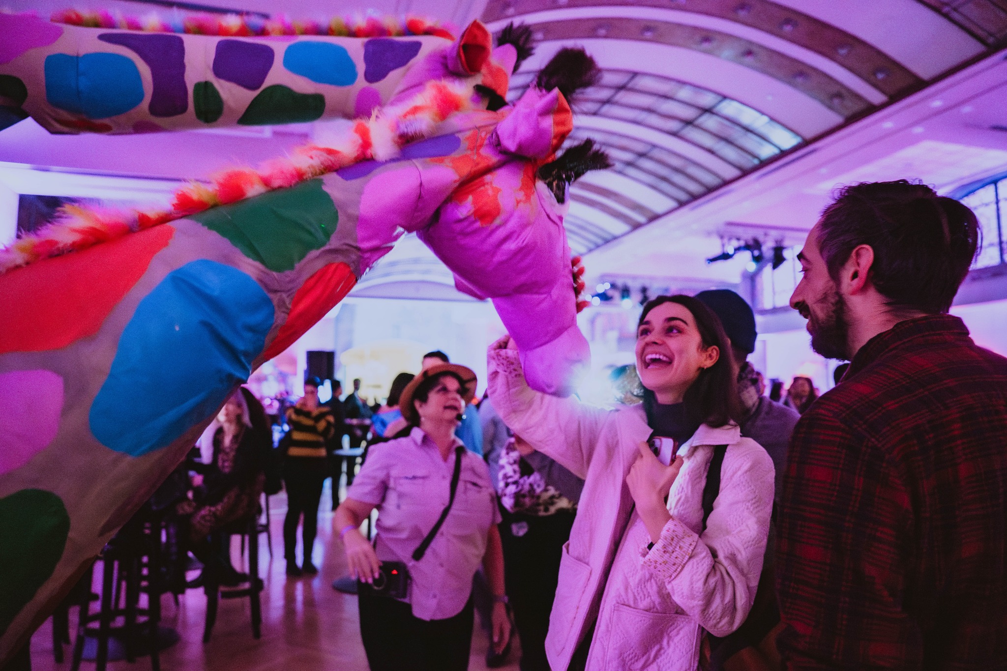 Une femme souriante touche le nez d'une grande marionnette girafe colorée à l'intérieur d'un grand espace intérieur voûté.