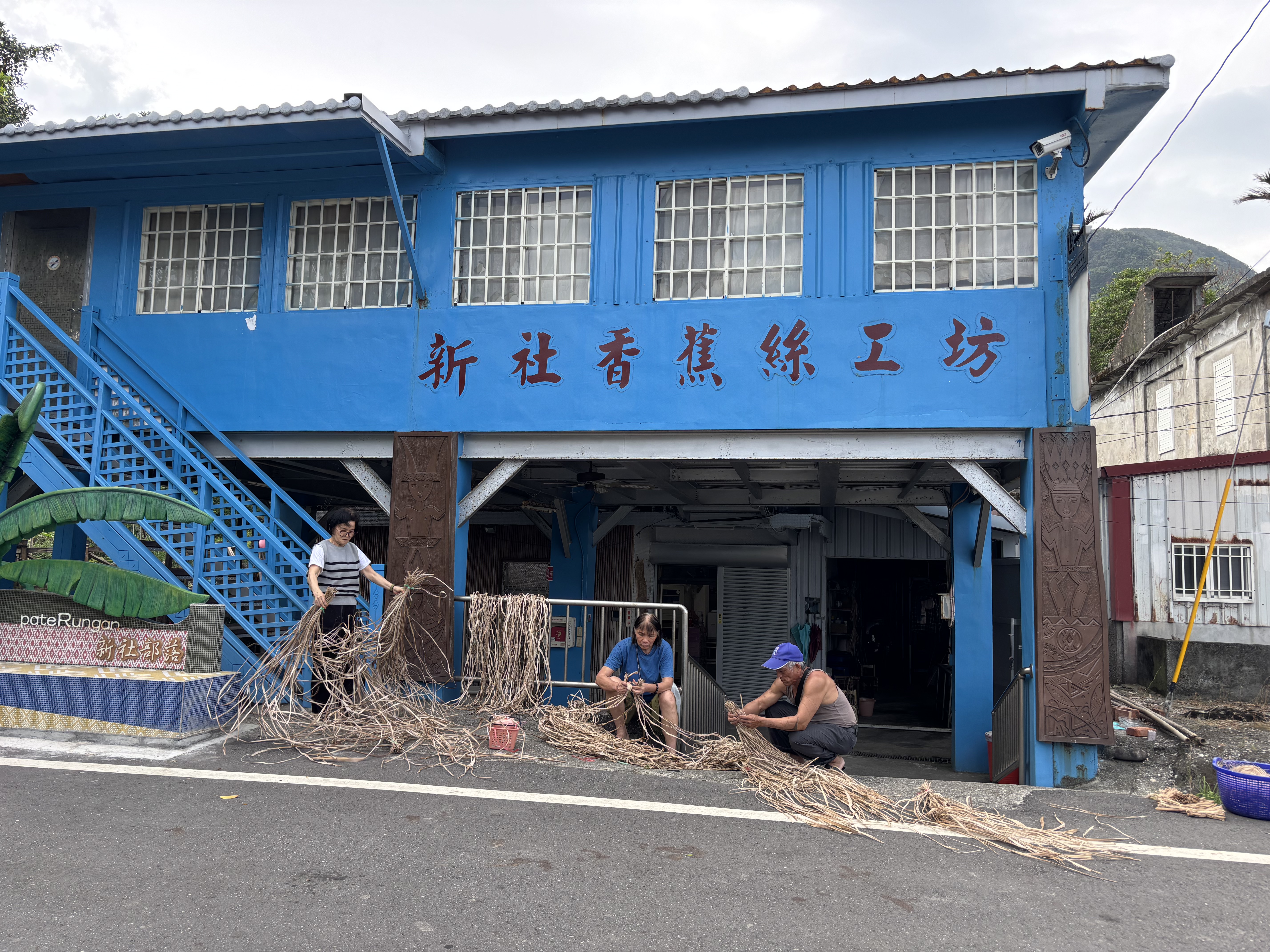 Three people sit outside a blue building sorting and weaving bundles of dried plant fibers along the roadside.