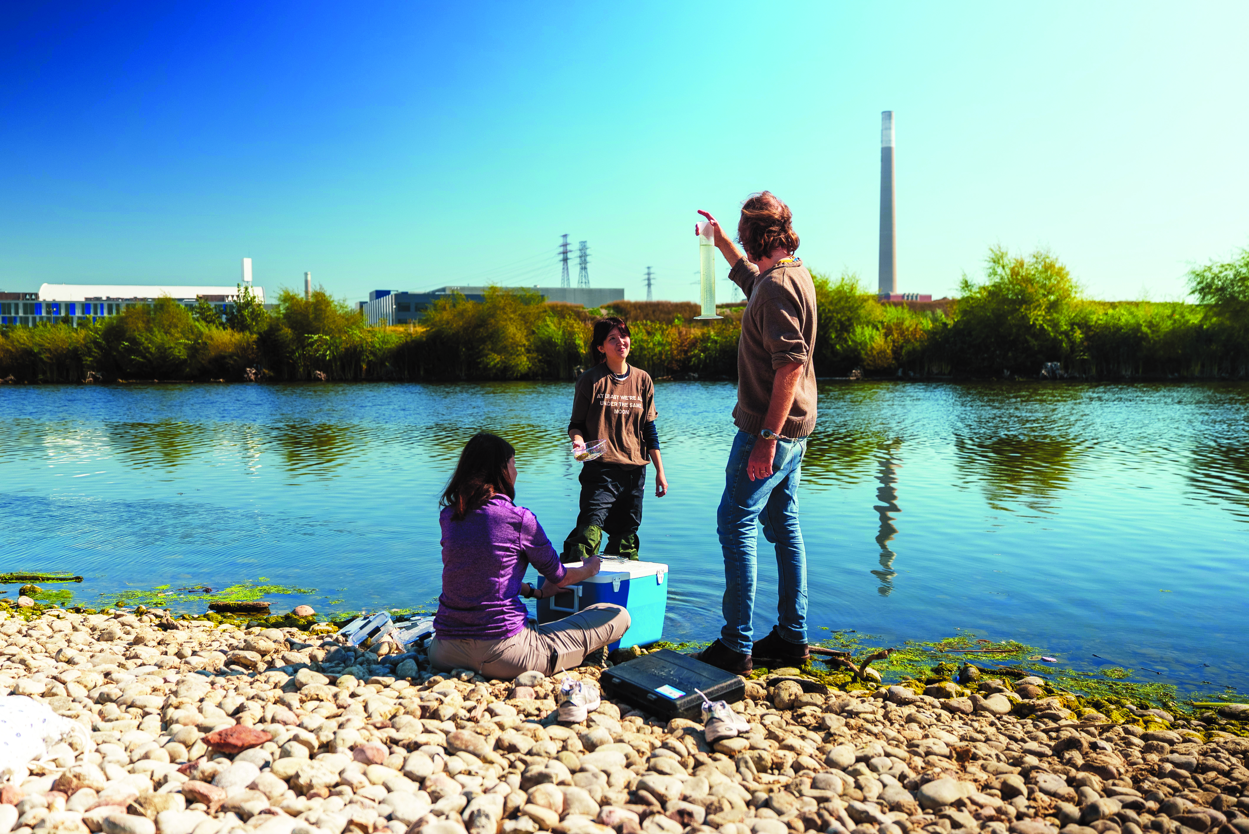 Three people conduct water sampling along a rocky shoreline, with one person holding up a clear measuring tube while others handle containers and equipment beside a river; trees, industrial buildings, and tall smokestacks are visible across the water under a clear blue sky.