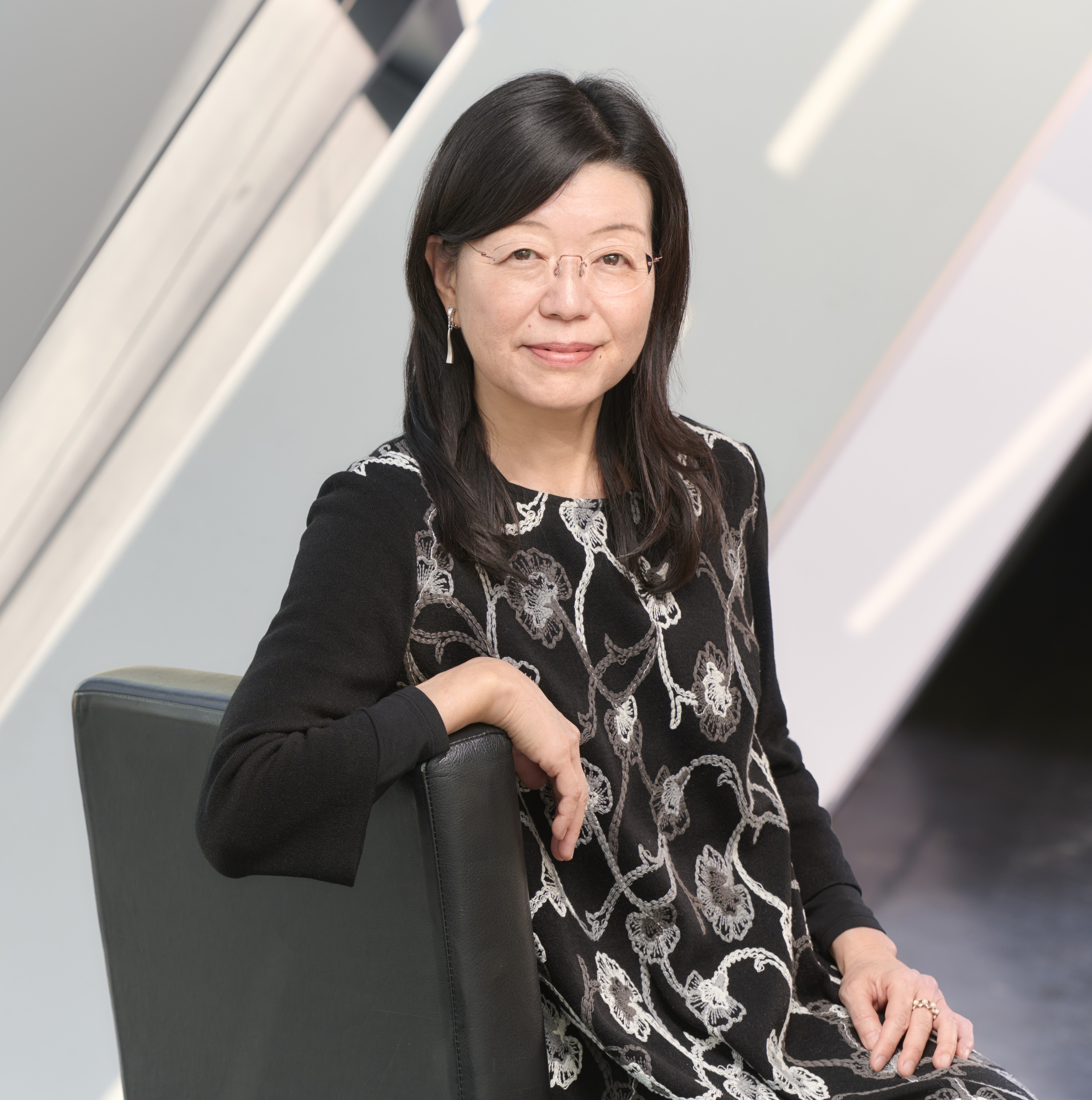 Asian woman with glasses wearing a black dress with a white floral pattern, sitting on a black chair against a modern background.