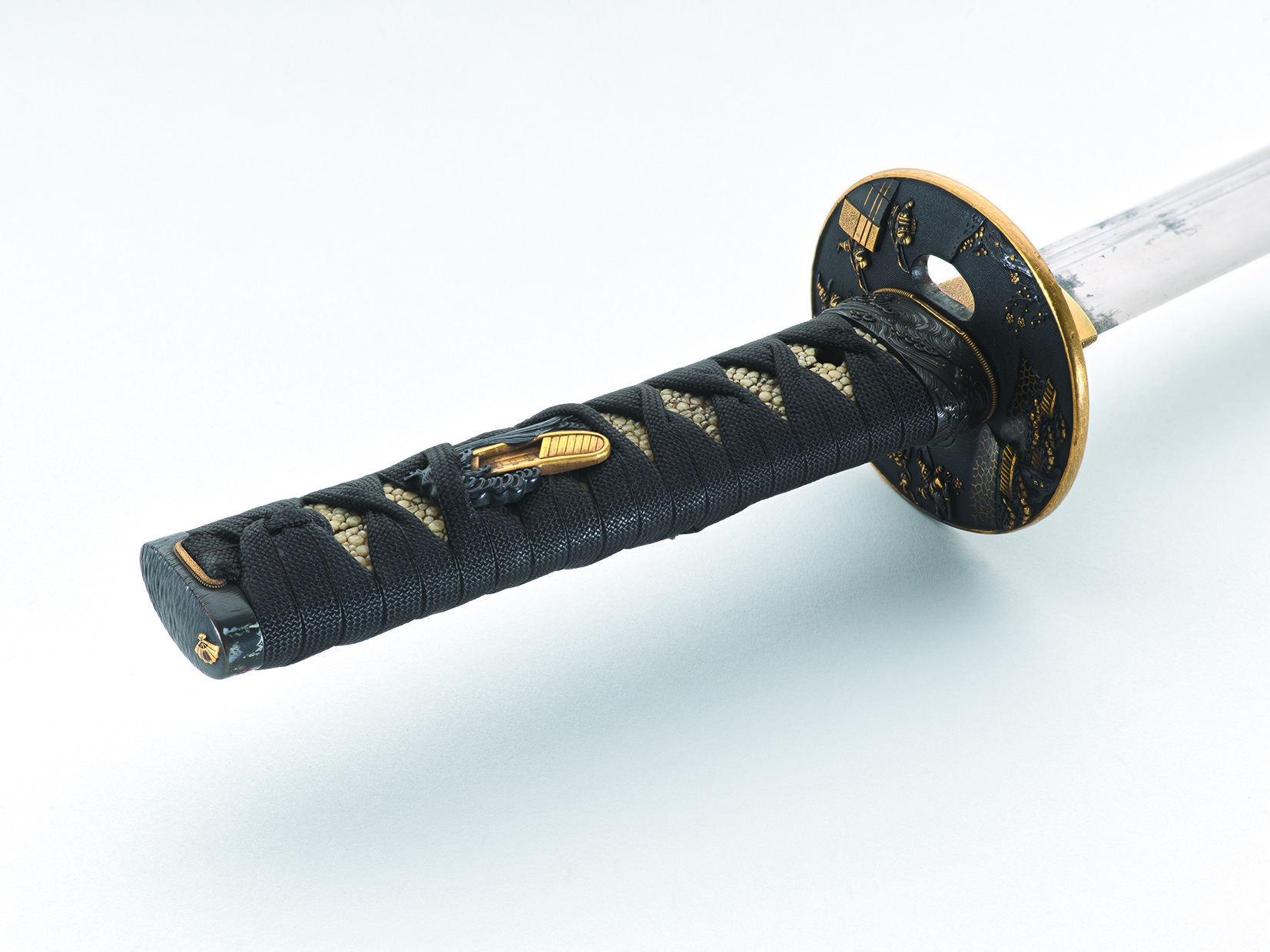 Close-up view of a Japanese sword hilt with black cord-wrapped handle, textured white ray-skin beneath, and ornate black-and-gold guard, photographed diagonally on a white background with soft lighting.