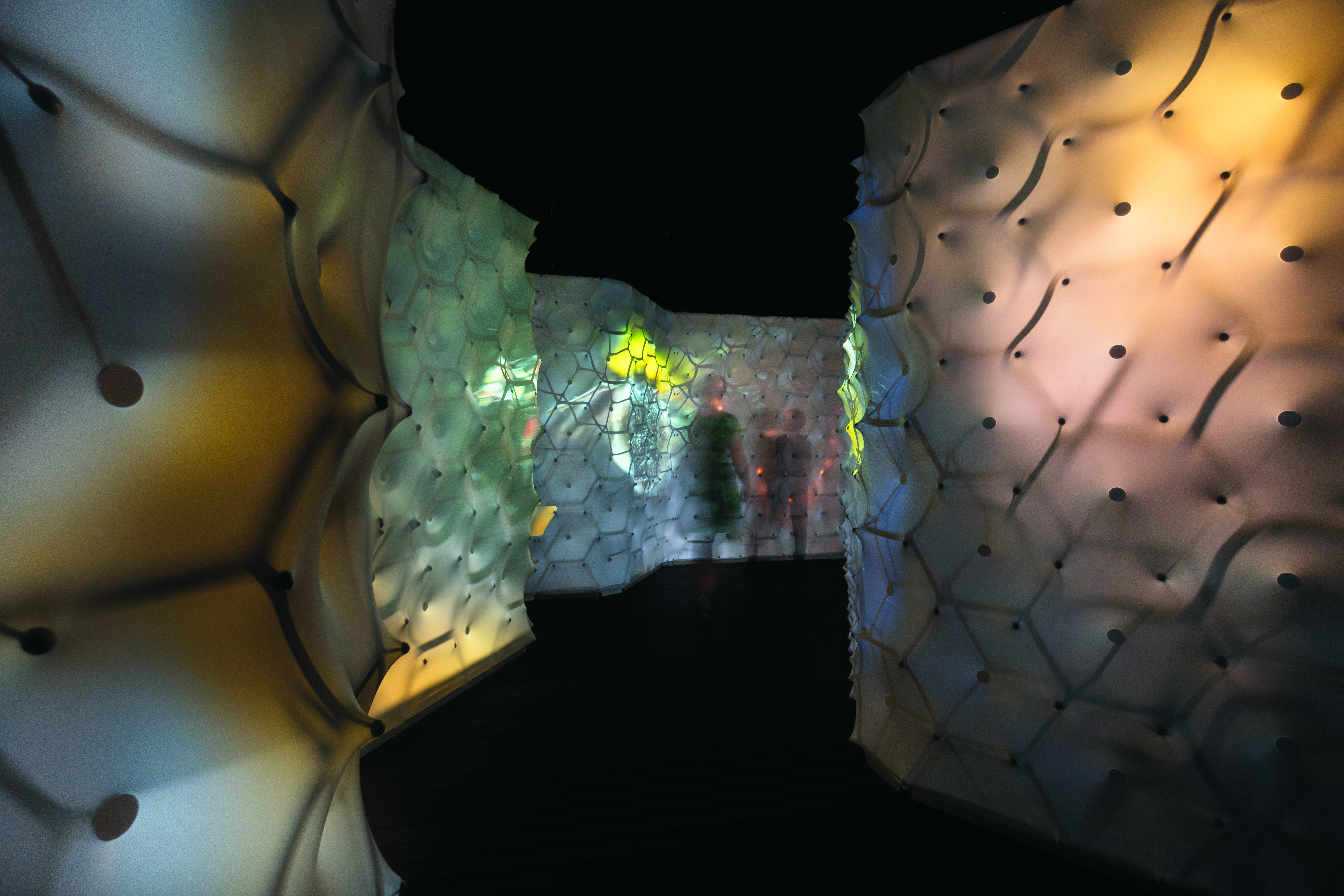 Espace d'installation immersif avec des murs en forme de nid d'abeille, doucement éclairés, formant un tunnel, vu de l'intérieur ; les surfaces translucides brillent dans des tons ambrés chauds, évoquant l'intérieur d'une ruche. 
