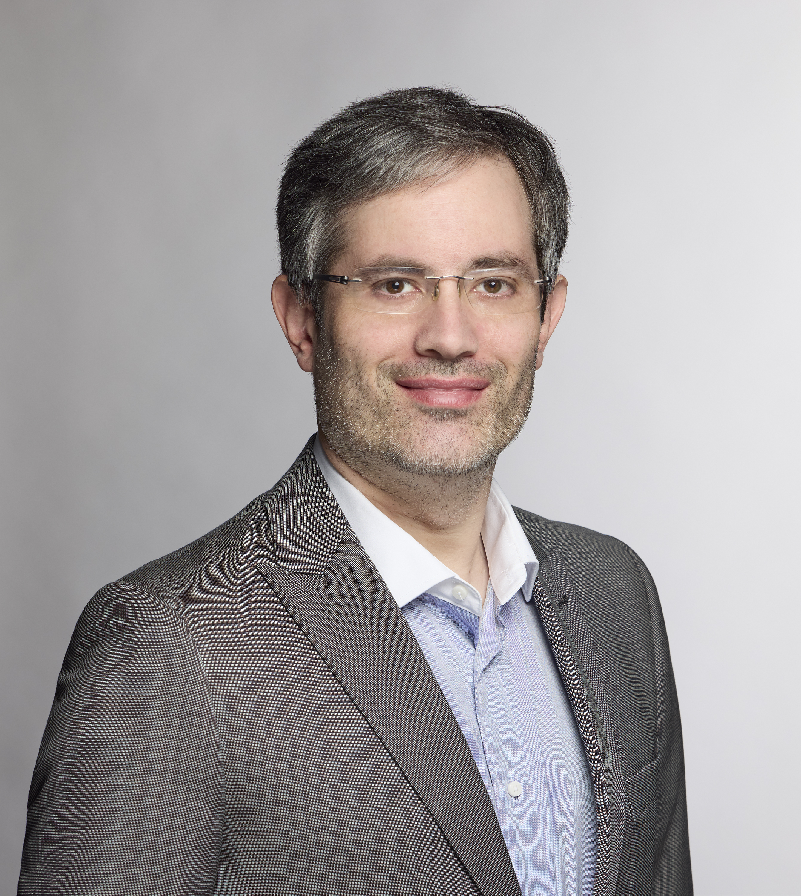 Portrait of a man with gray hair and glasses wearing a gray suit jacket over a light blue shirt against a plain light gray background.