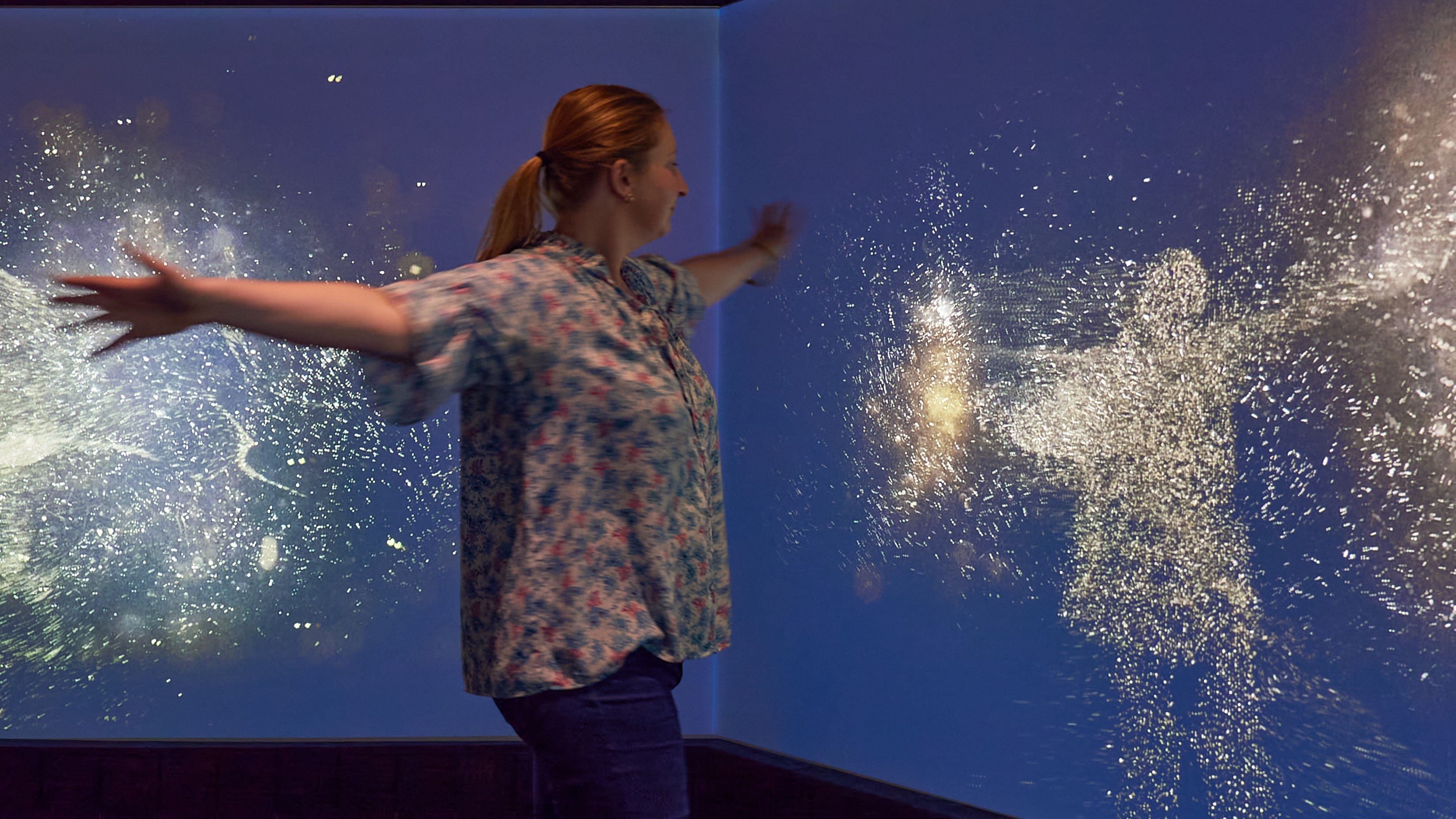 A woman moves in front of an interactive screen where clusters of pollen-like light follow her movements.