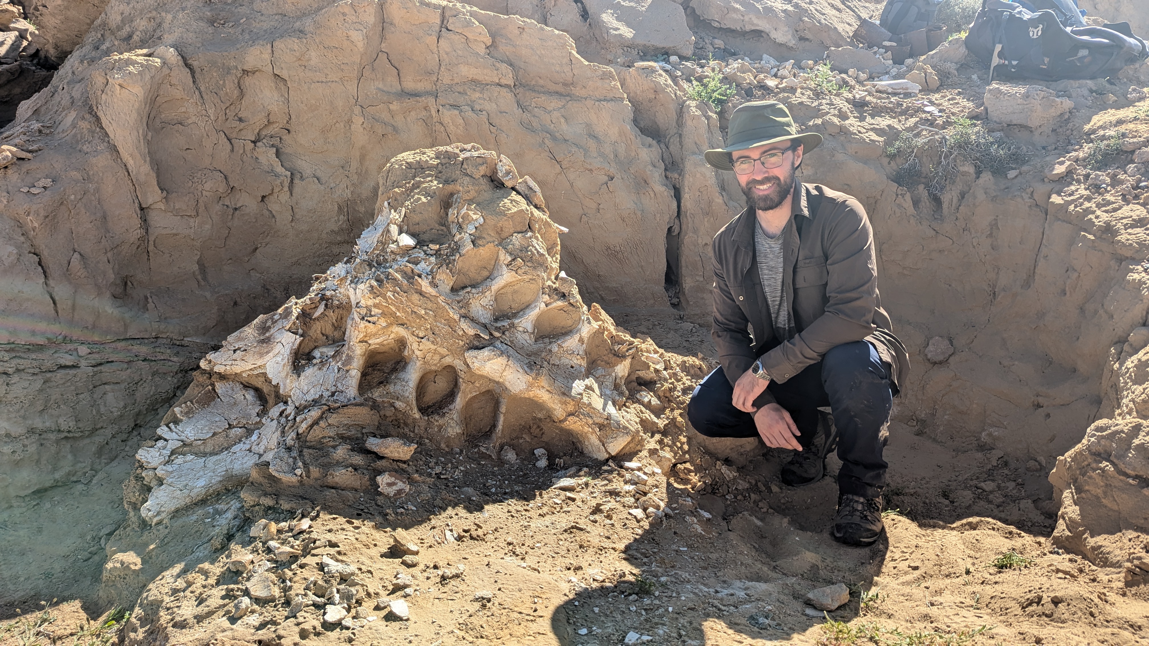 Tom Dudgeson wearing a baseball cap, sunglasses, and dark outdoor clothing crouches beside a large dinosaur skull partially embedded in rocky terrain.