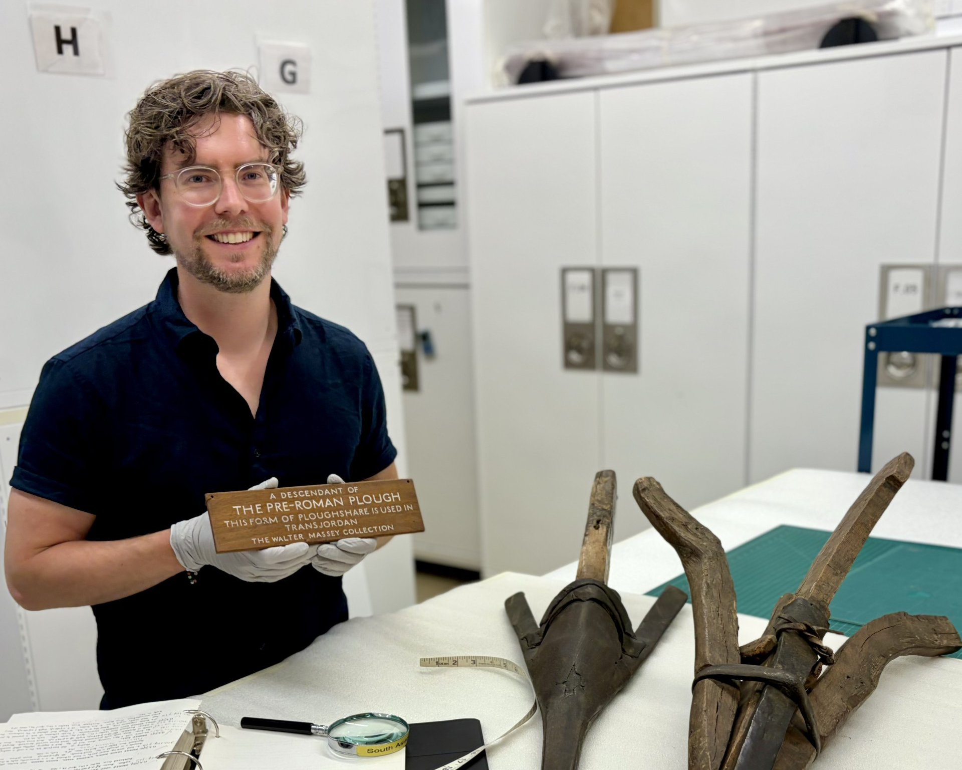 Greg Fester holding a wooden label that reads "MANITOBA - Plains Indian hide scraper," with three large hide scrapers on a table.