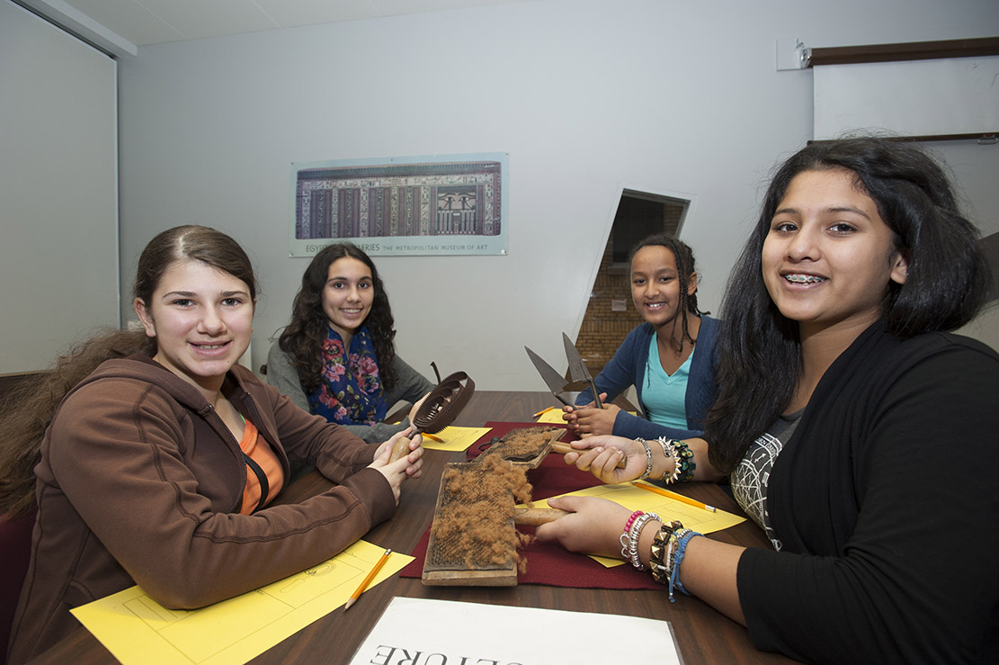 Four smiling students are looking at the camera sitting around a table holding a different tool.  One is holding a curry comb, one holds carding combs, and one is holding sheep shears.  