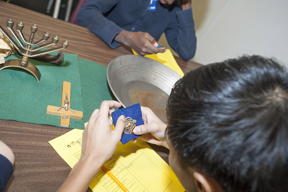 A student is holding a pin, while on the table in front of them is a menorah, a crucifix, and a tray for panning for gold. 