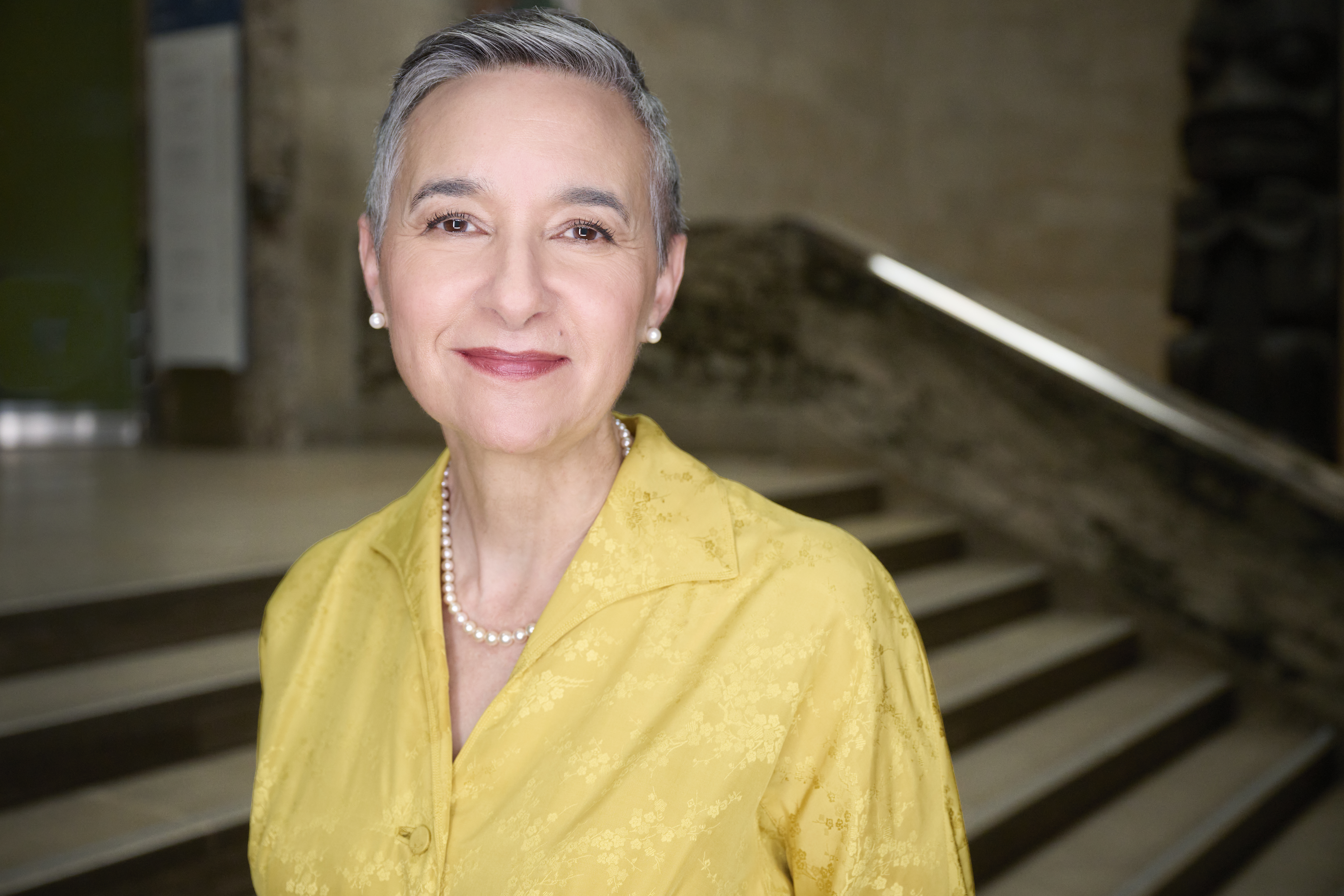 Portrait of a smiling person with short gray hair wearing a yellow blouse and pearl necklace, standing indoors near stairs.