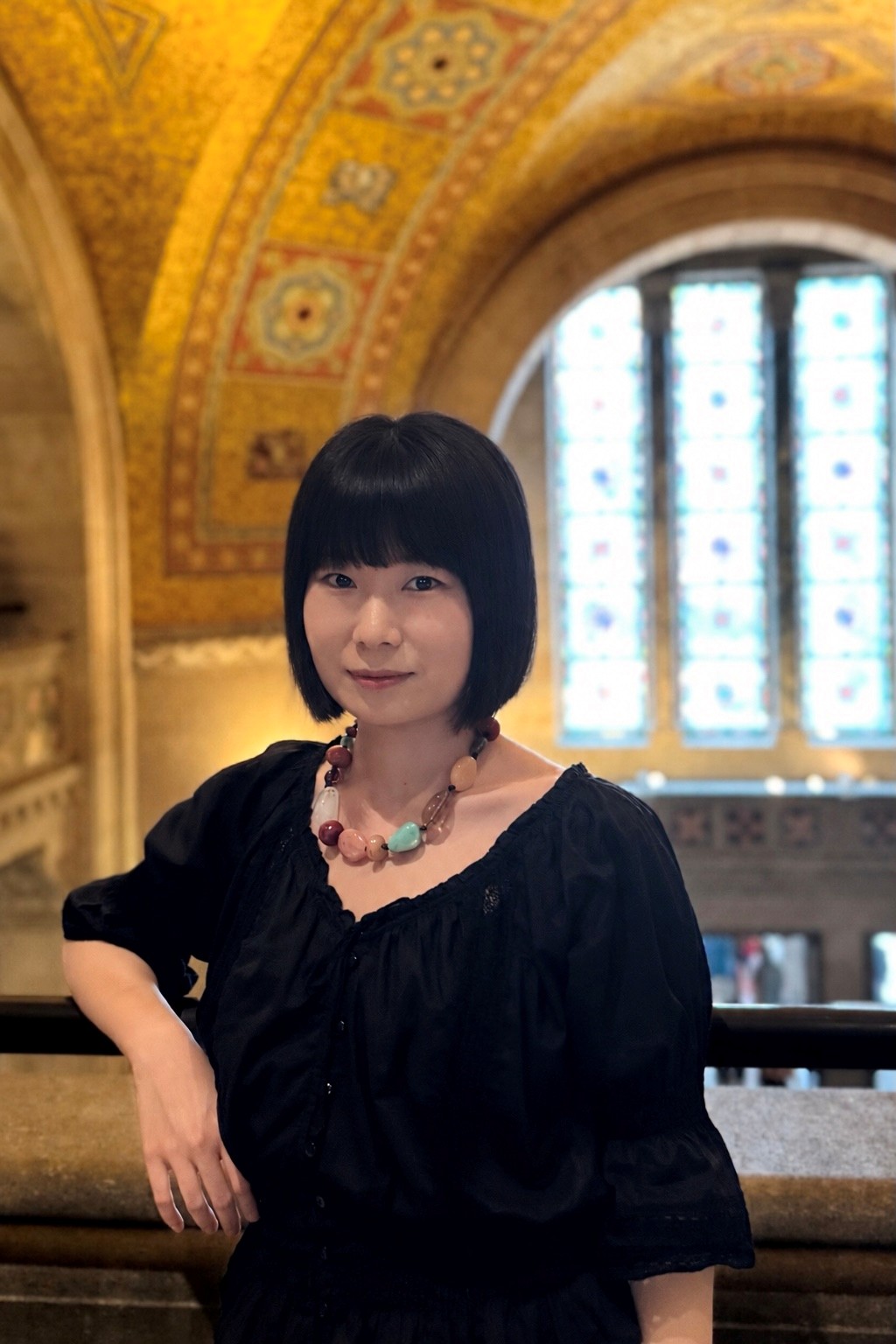 Portrait of a woman with short black hair, wearing a black blouse and necklace, standing indoors with an arched window and ornate ceiling in the background.