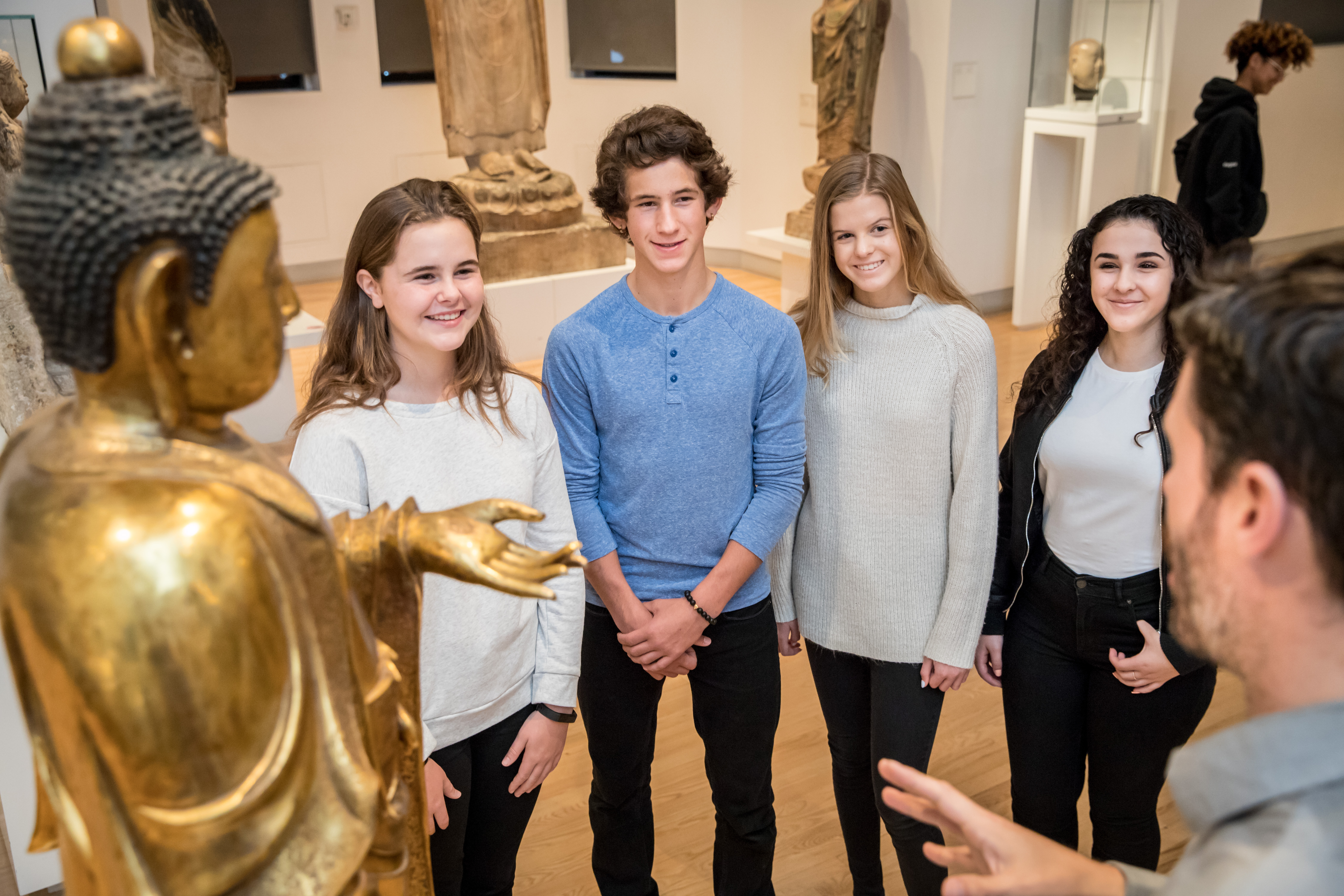 Four smiling students listen intently to a Museum Educator telling them about a golden Buddha statue.