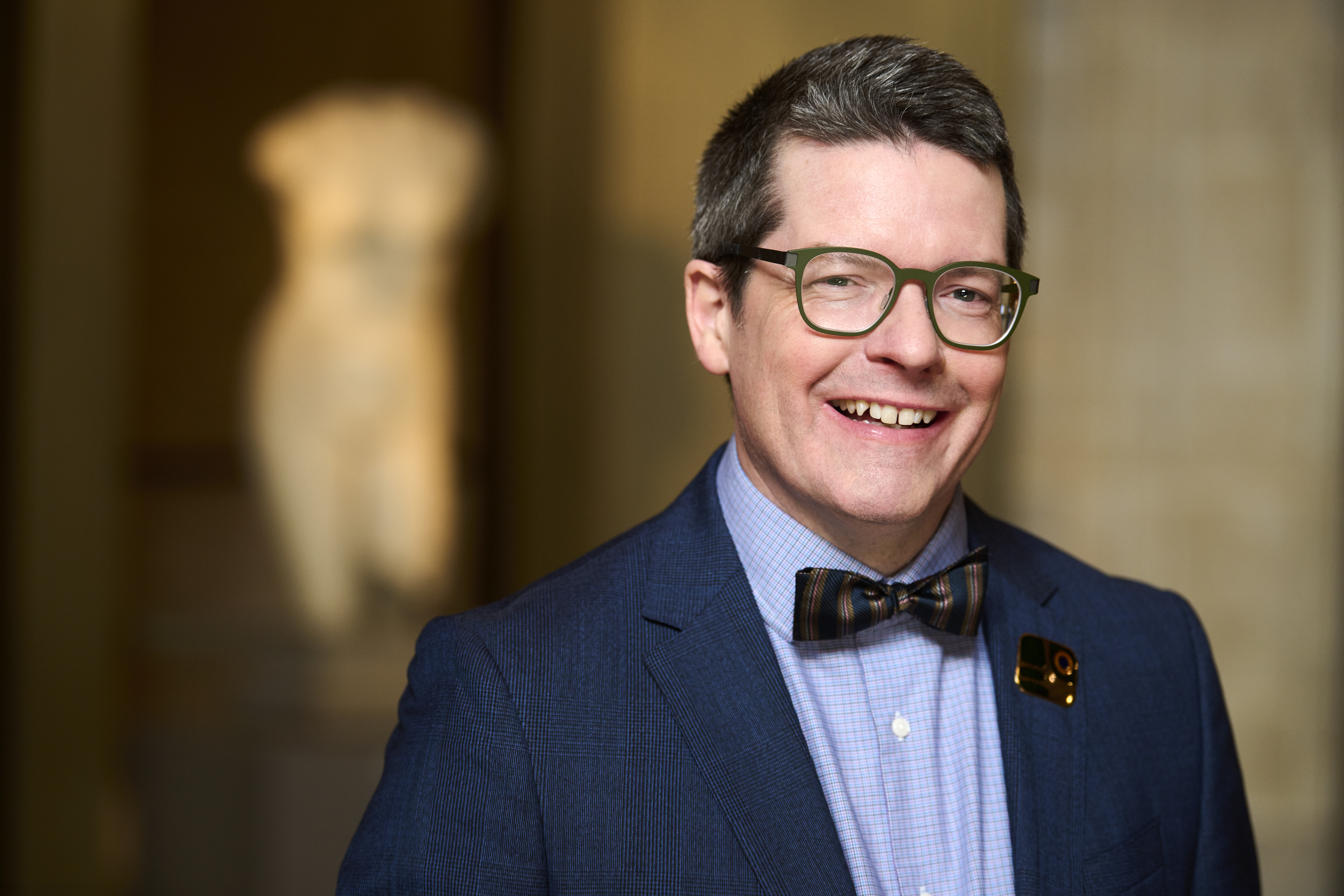 Portrait of Nicholas Bell wearing glasses, a navy suit, bow tie, and a floral lapel pin, with a blurred museum artifact in the background.