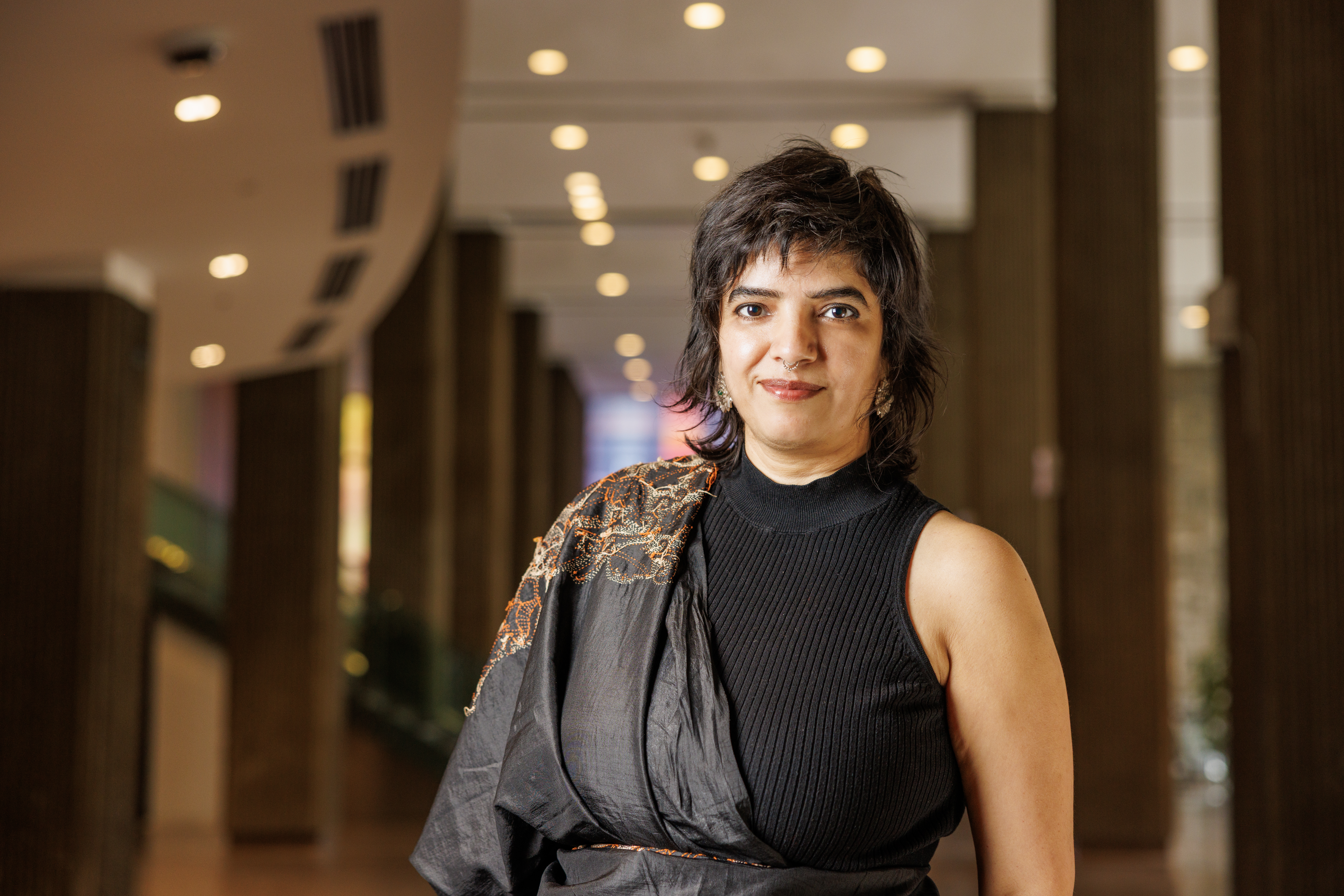 Portrait of Pramila Choudhary standing indoors with a blurred modern architectural background.