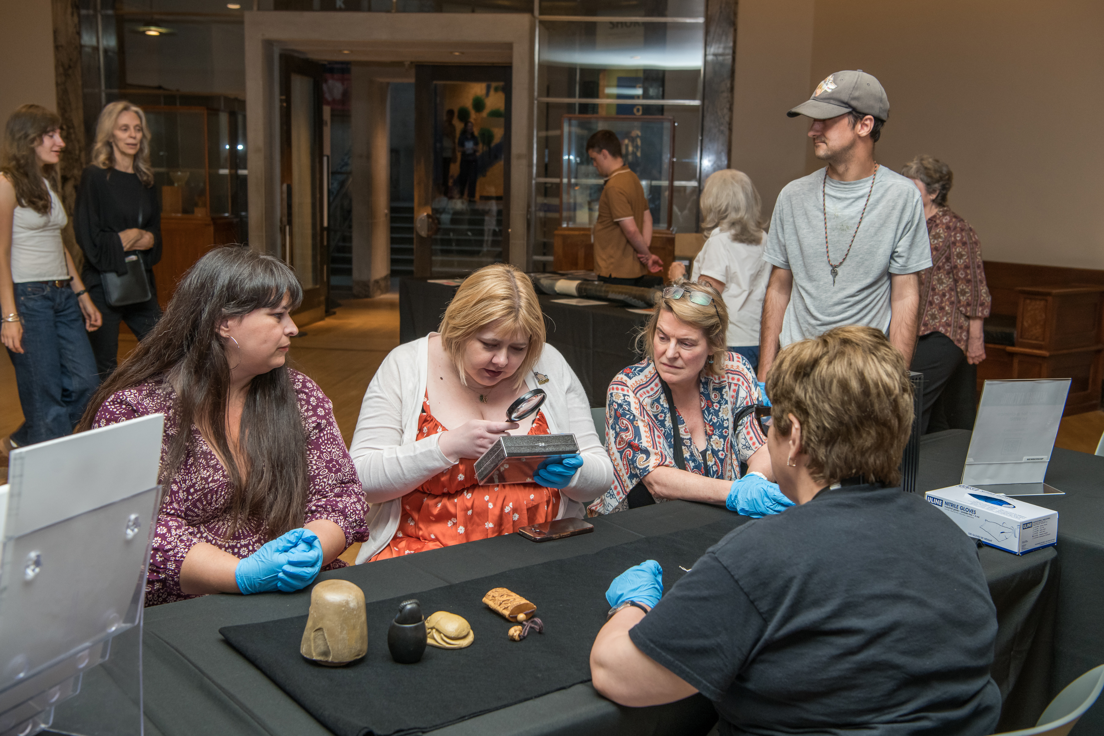 Four people seated at a table examining fossilized skulls, with visitors and a museum entrance in the background.
